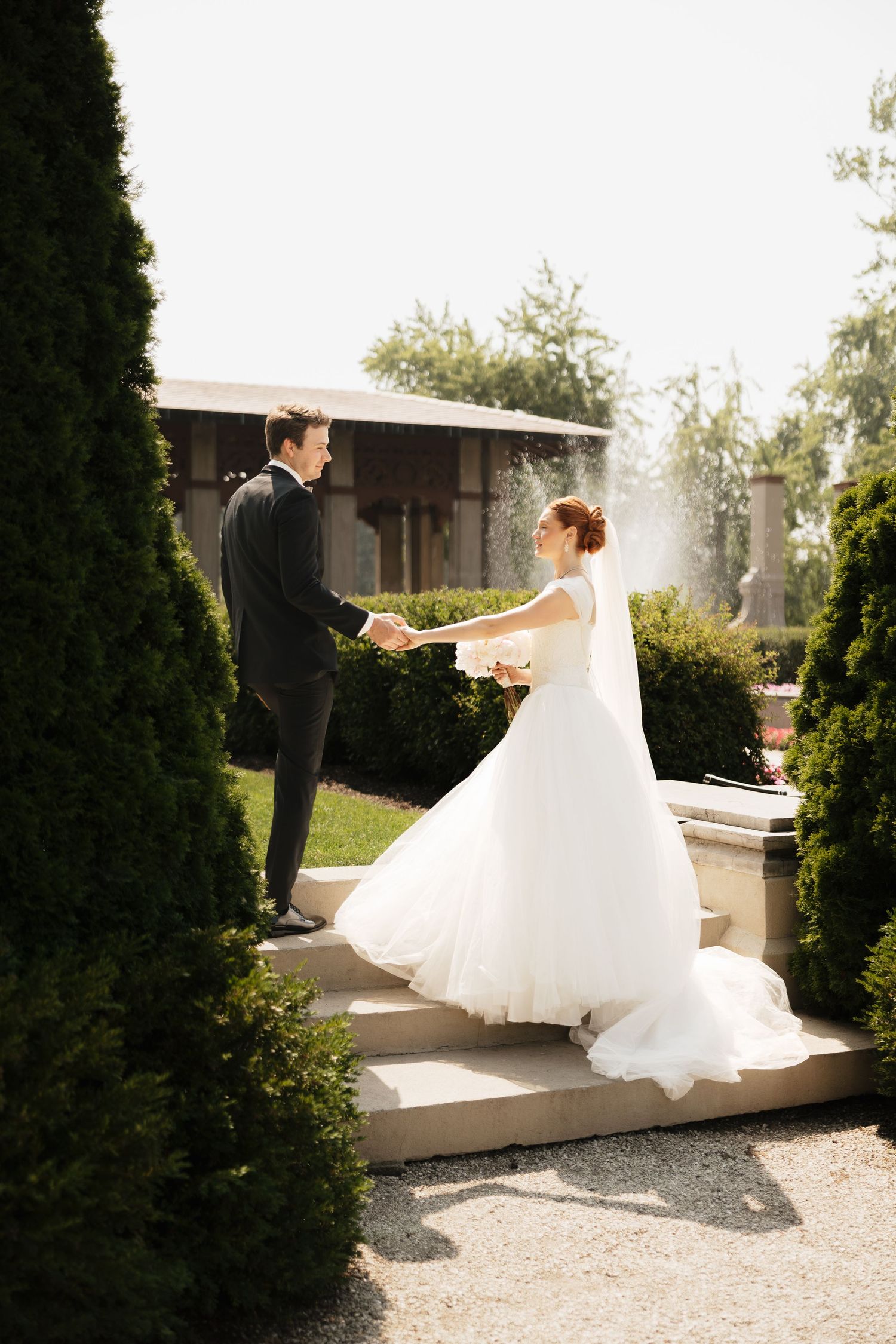 Bride in white gown and groom in black suit hold hands on garden steps surrounded by lush greenery.