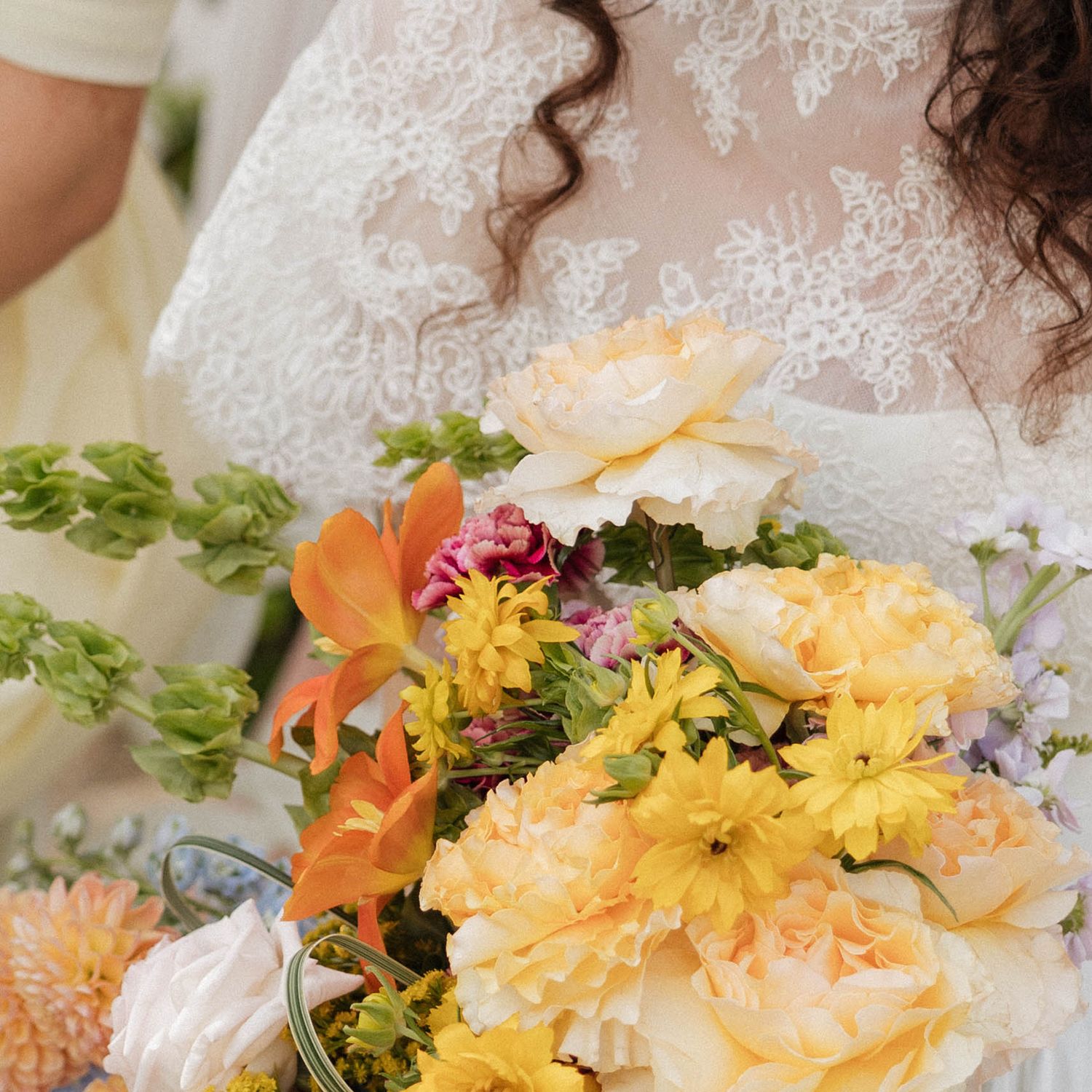 Bride in lace wedding dress holding a vibrant bouquet of yellow, peach, and orange flowers including peonies and dahlias.