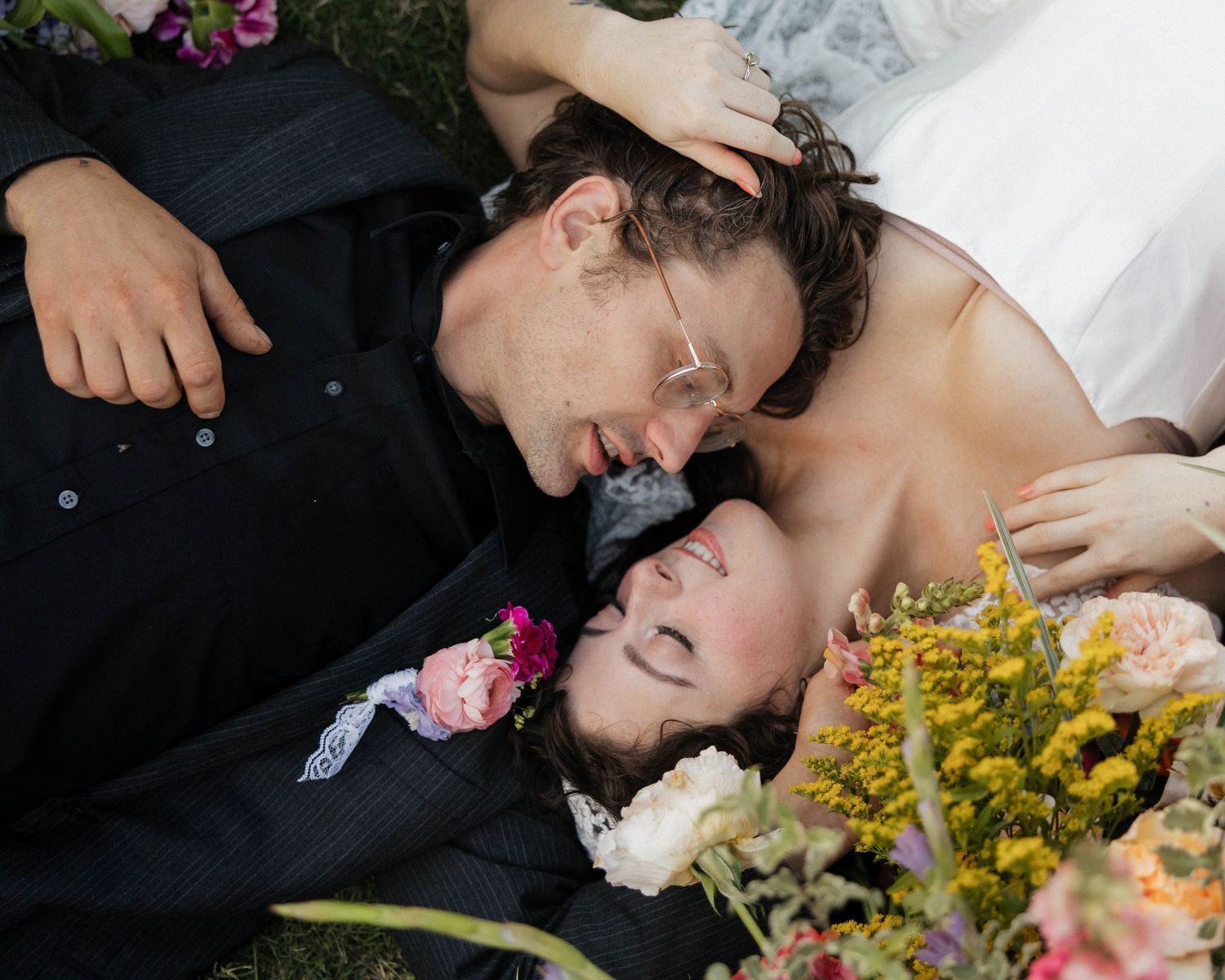 Bride and groom lying on grass surrounded by colorful flowers in a romantic wedding portrait.