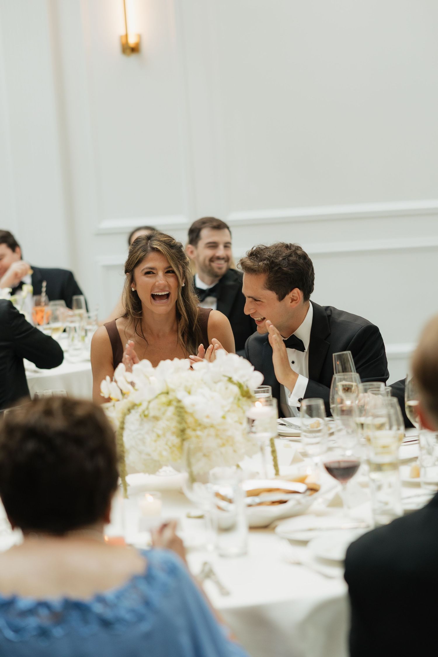 Bride and groom laughing joyfully at wedding reception dinner table with white floral centerpiece.