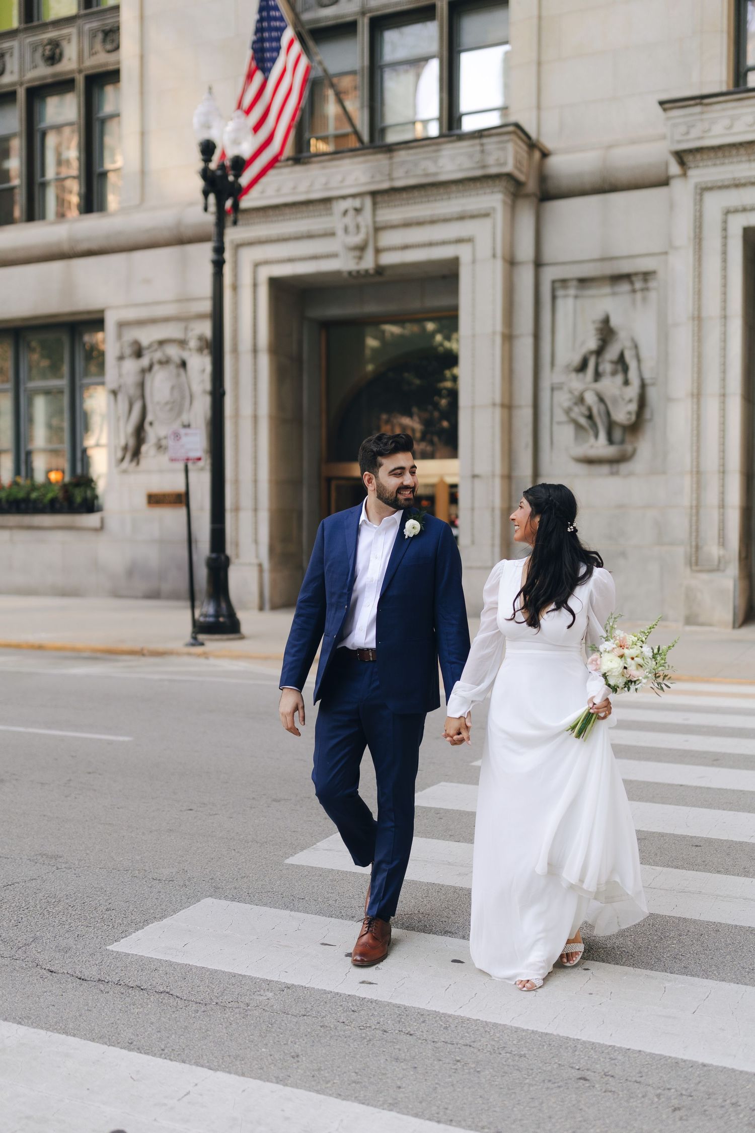 A couple in wedding attire walks together outside a grand courthouse building with an American flag flying above.