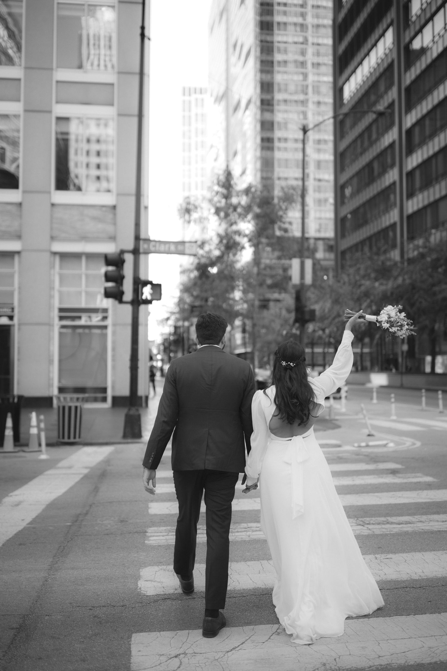 A newlywed couple walks away from camera on an urban sidewalk with modern buildings in black and white.