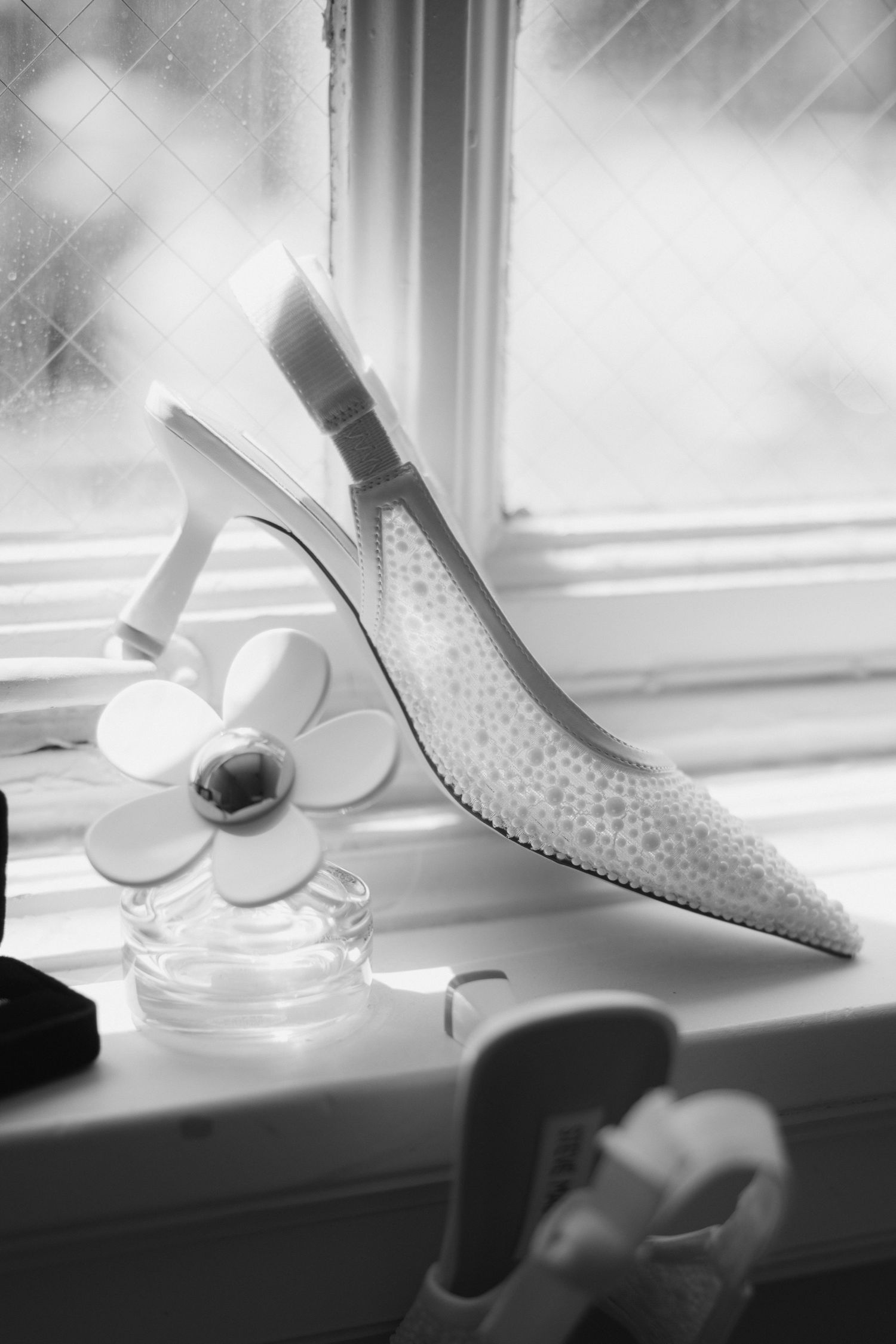 Black and white photo of a beaded bridal heel shoe on a windowsill beside a floral perfume bottle.