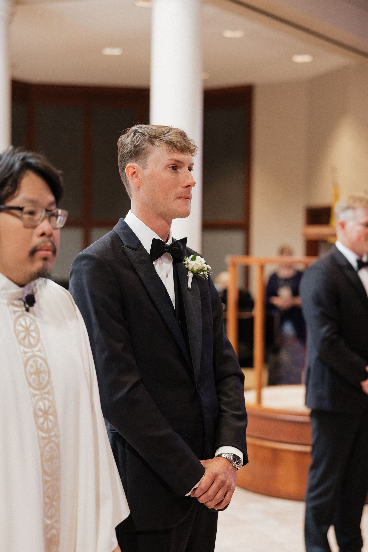 Groom in black tuxedo with boutonniere stands at church altar beside officiant during wedding ceremony.