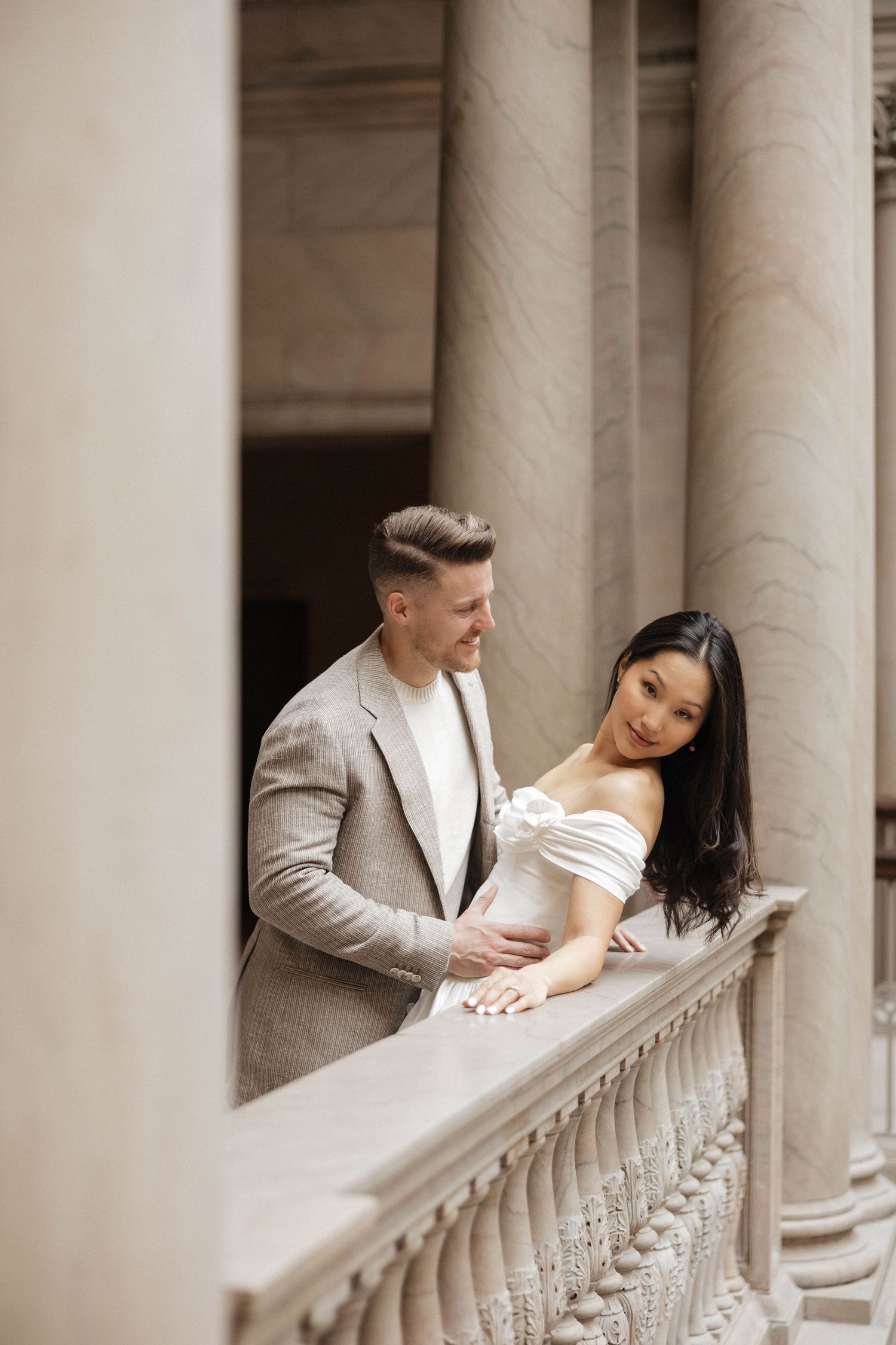 Romantic couple standing together on an ornate stone balustrade surrounded by towering marble columns.