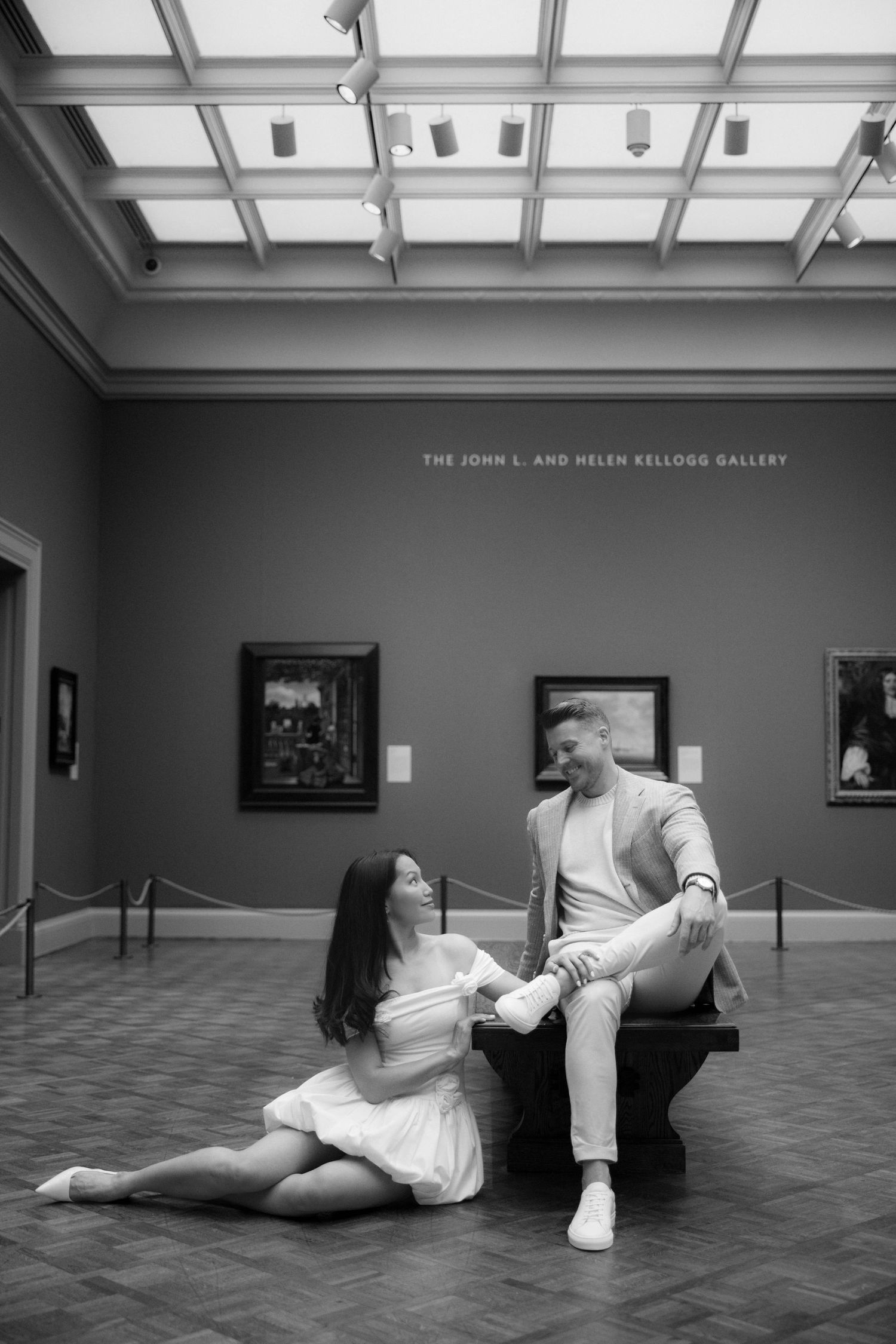 Couple poses in black and white at the John L. and Helen Kellogg Gallery, woman sitting on floor, man on bench.