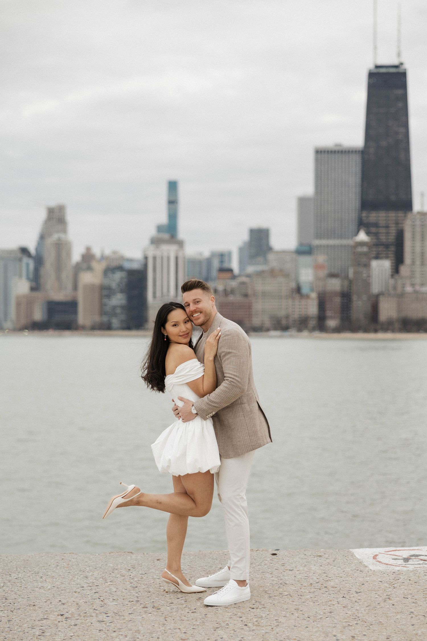 Couple embracing on Chicago lakefront with city skyline backdrop, woman in white dress, man in tan blazer.