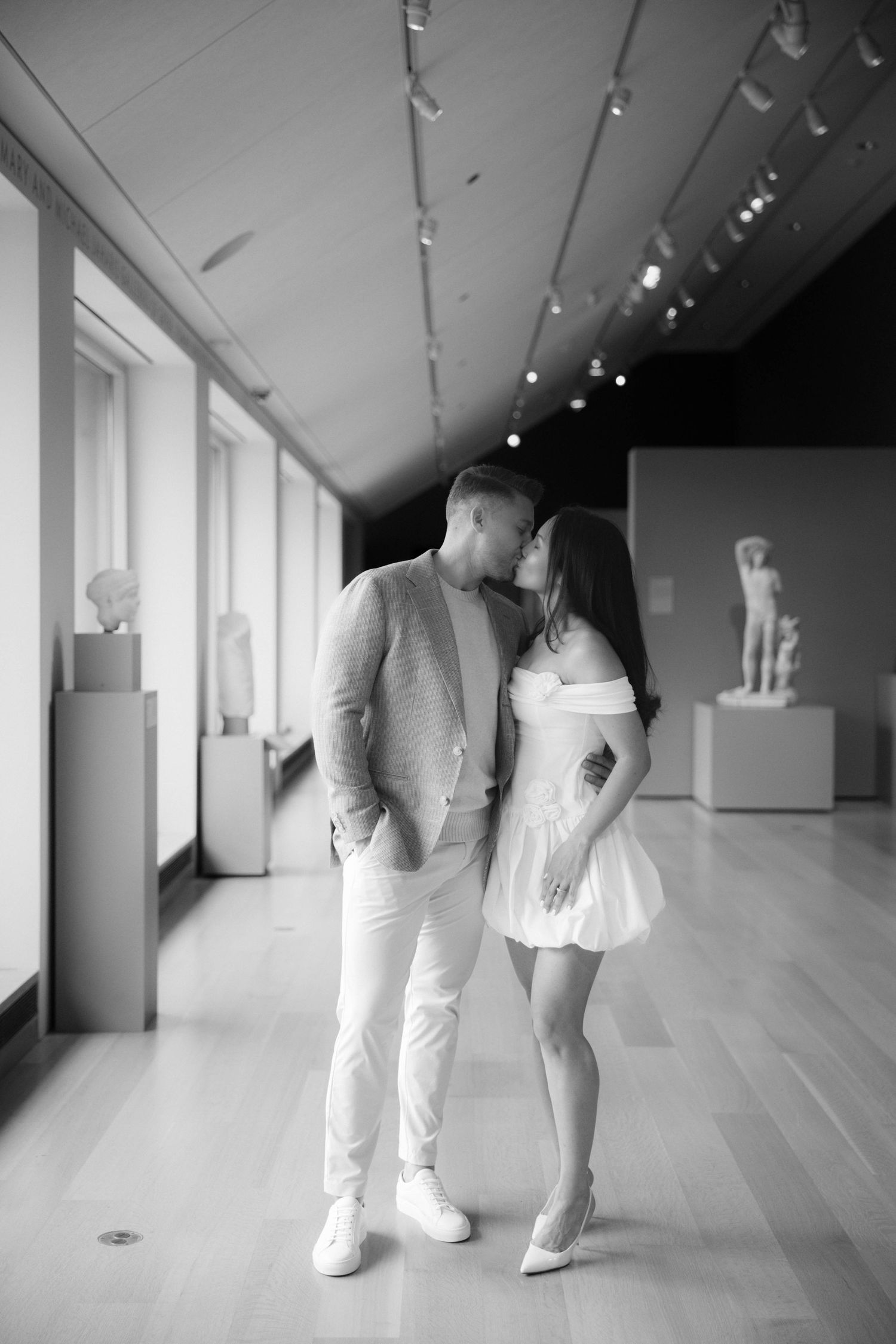 Black and white photo of a couple sharing a kiss in an art museum gallery with classical sculptures.