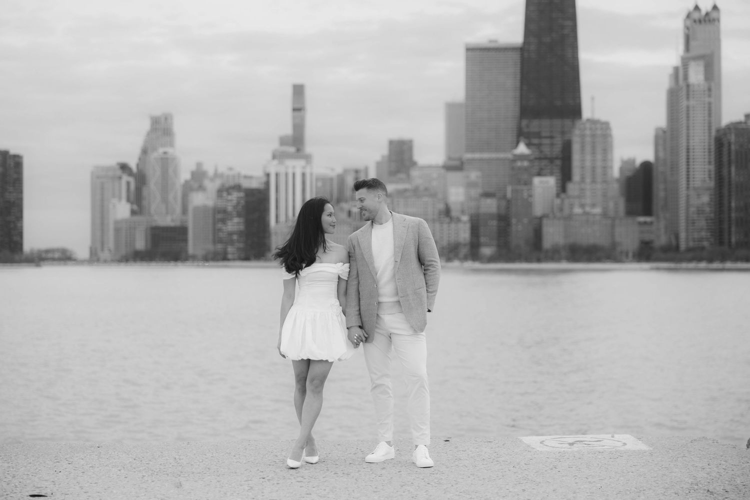 Couple in white attire sharing a romantic moment with the Chicago city skyline in black and white photo.