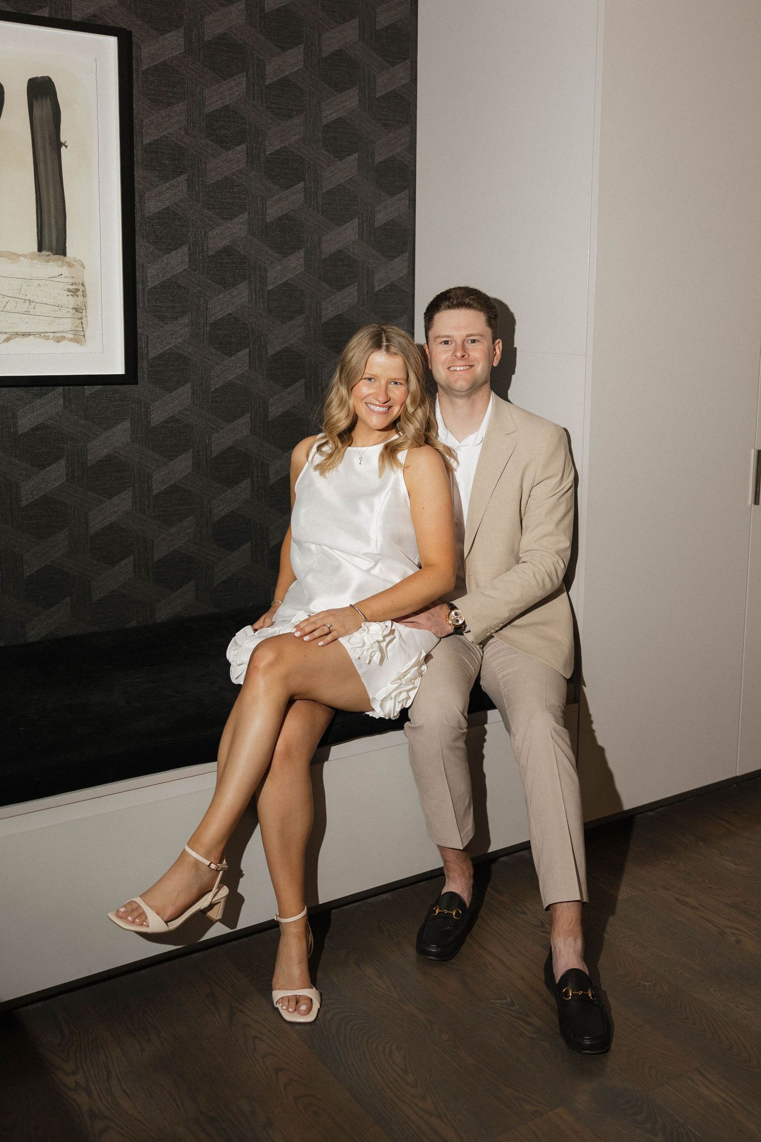 Couple sitting together indoors on bench, woman in white dress and man in beige suit, smiling.