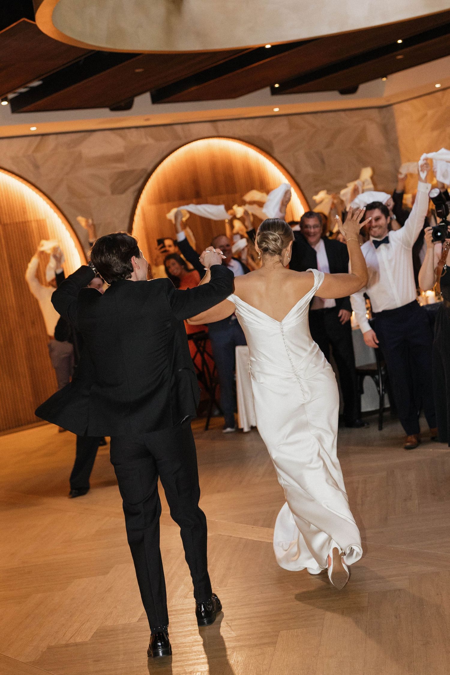 Bride and groom make grand entrance onto dance floor as wedding guests cheer and wave napkins in elegant venue.