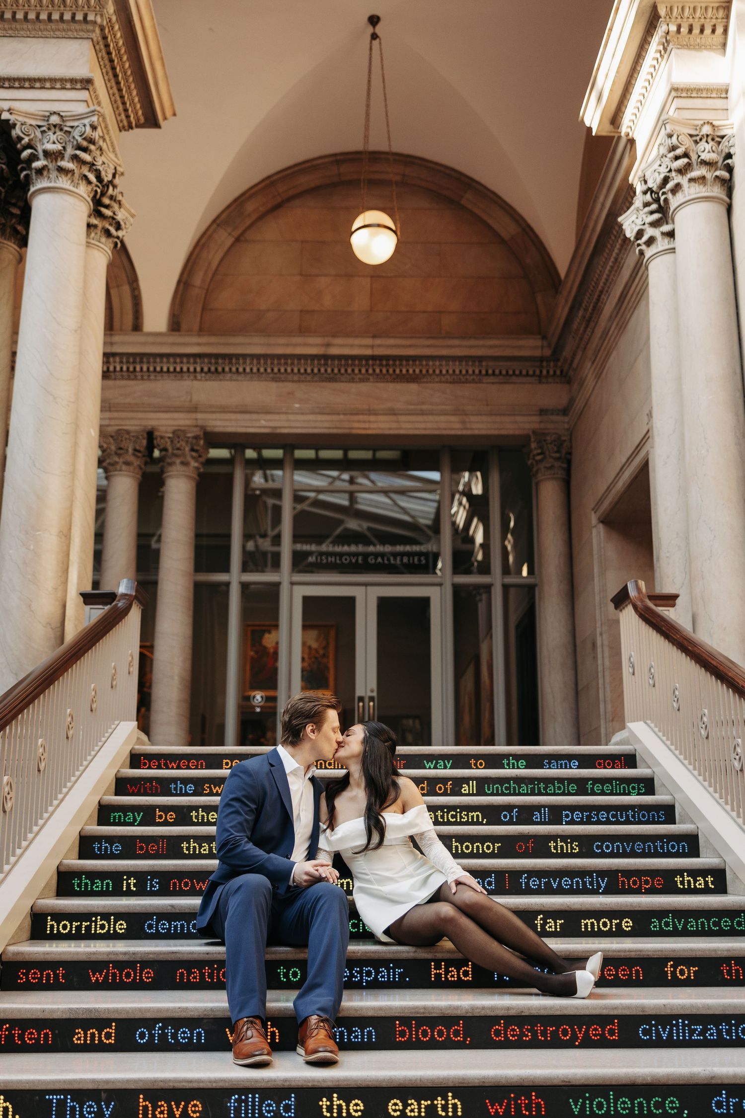 Couple sitting on steps in an elegant building interior with classical architecture and columns.