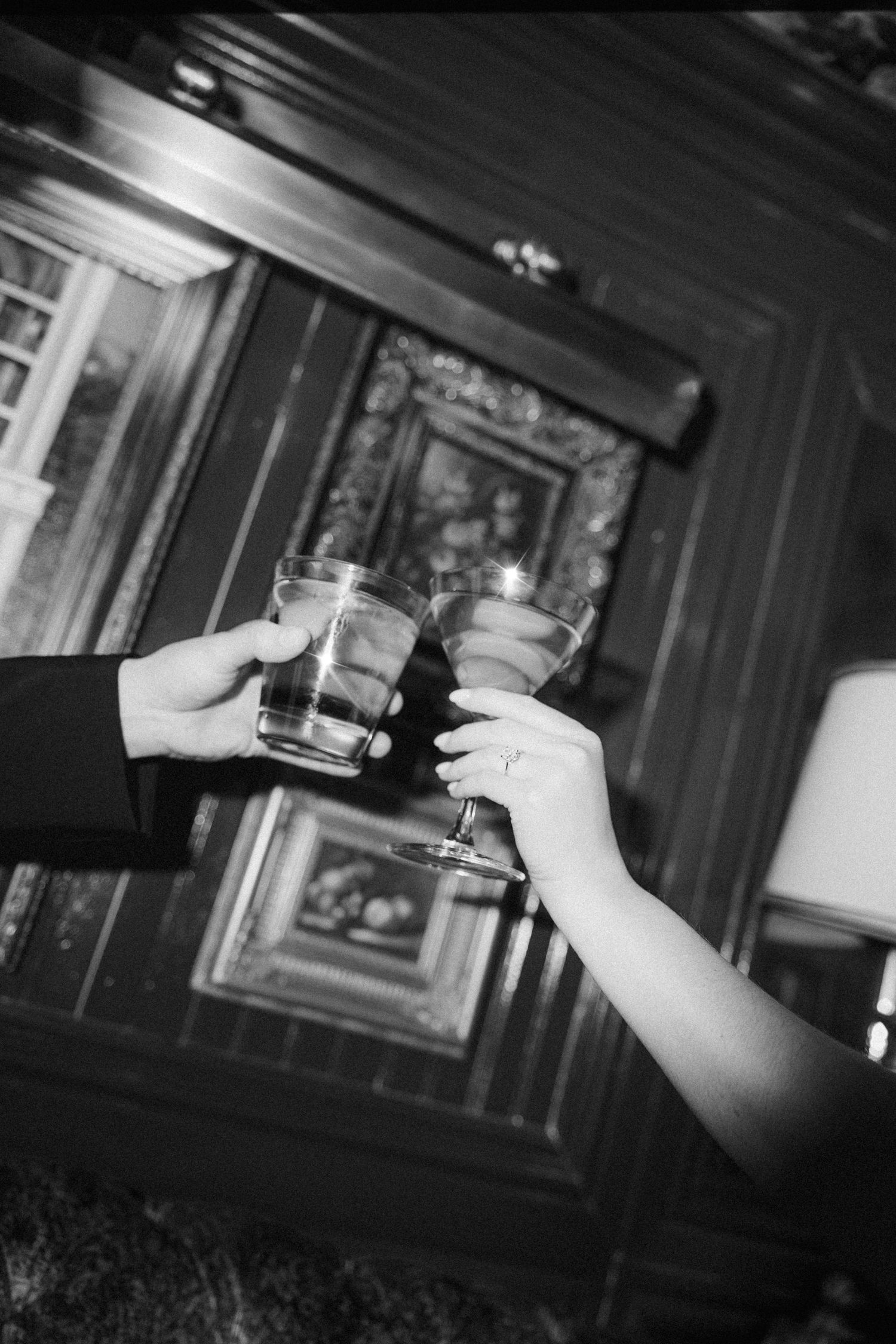 Two hands raise glasses together in a toast at a dimly lit bar with wood paneling and framed artwork in the background.