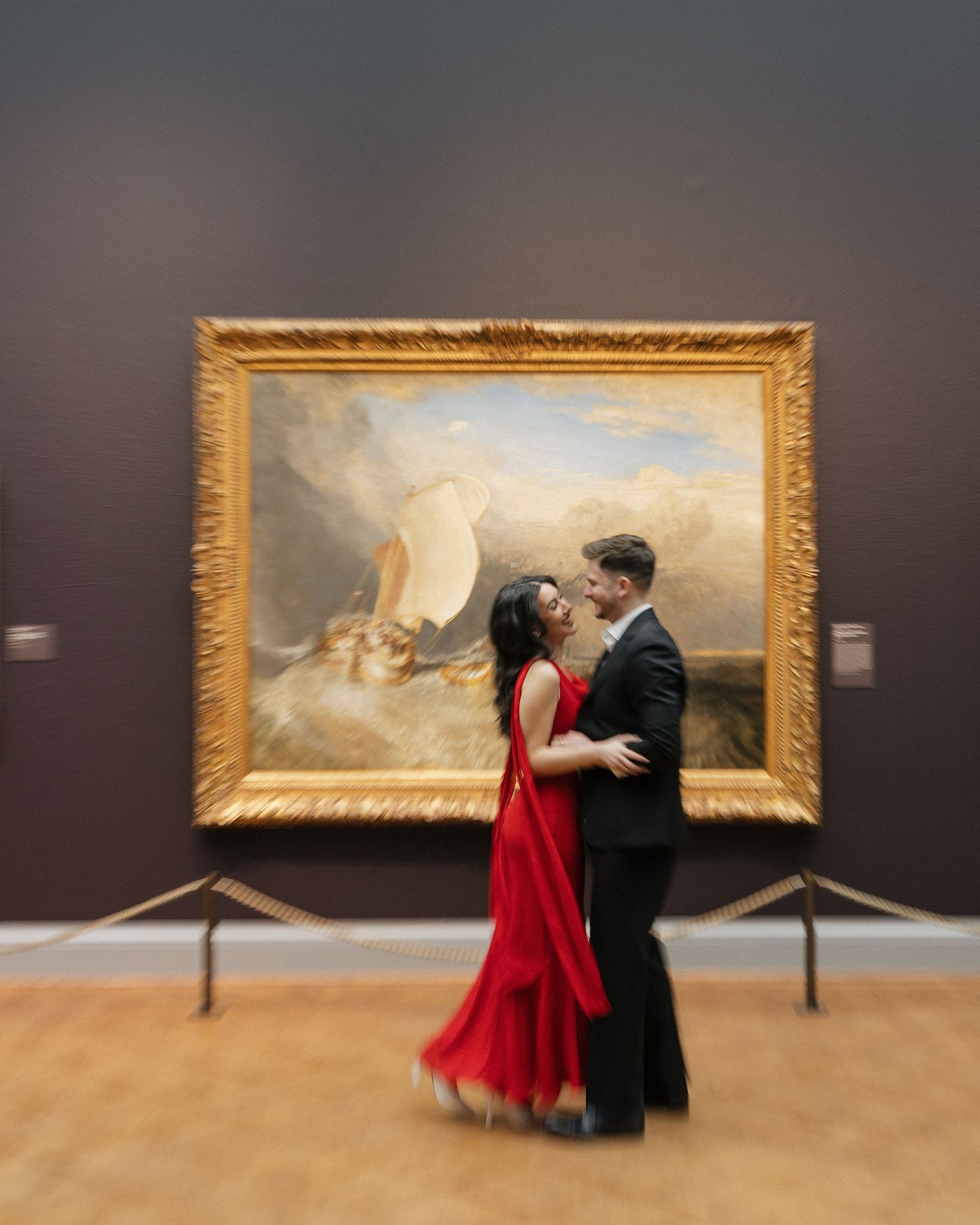 A couple dances together in a museum gallery in front of a large ornate gold-framed classical painting.