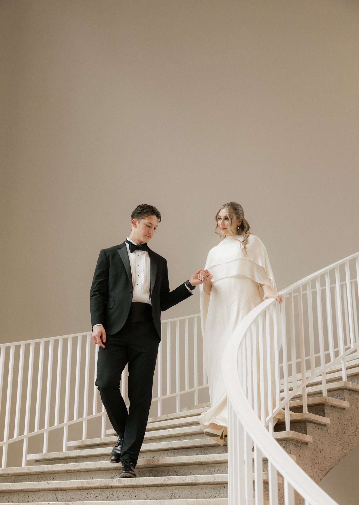 Couple in wedding attire poses on white staircase against neutral background.