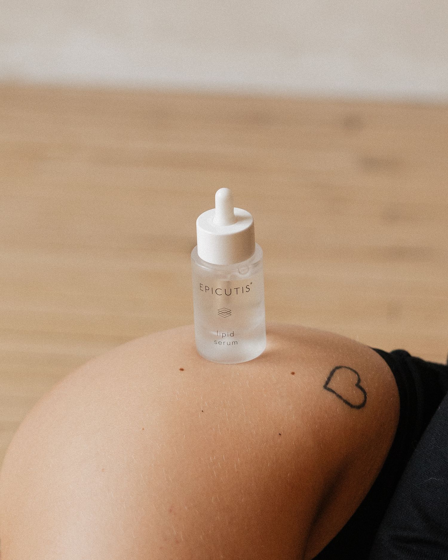 Close-up of Epicutis serum bottle balanced on skin with small heart tattoo visible.