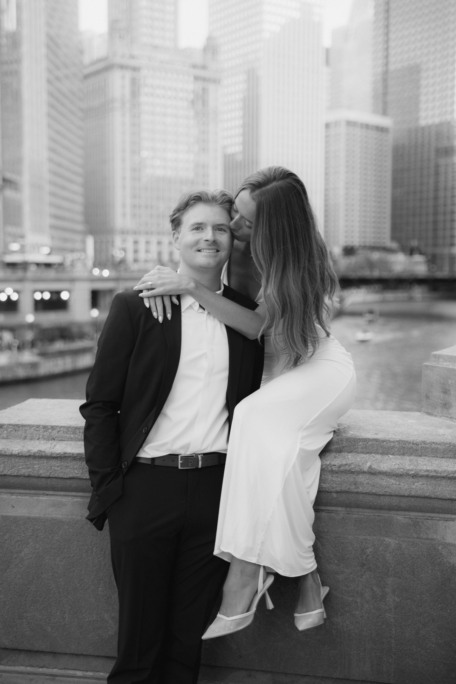 Black and white photo of a couple embracing in front of Chicago River skyline, woman in white dress kissing man in suit.