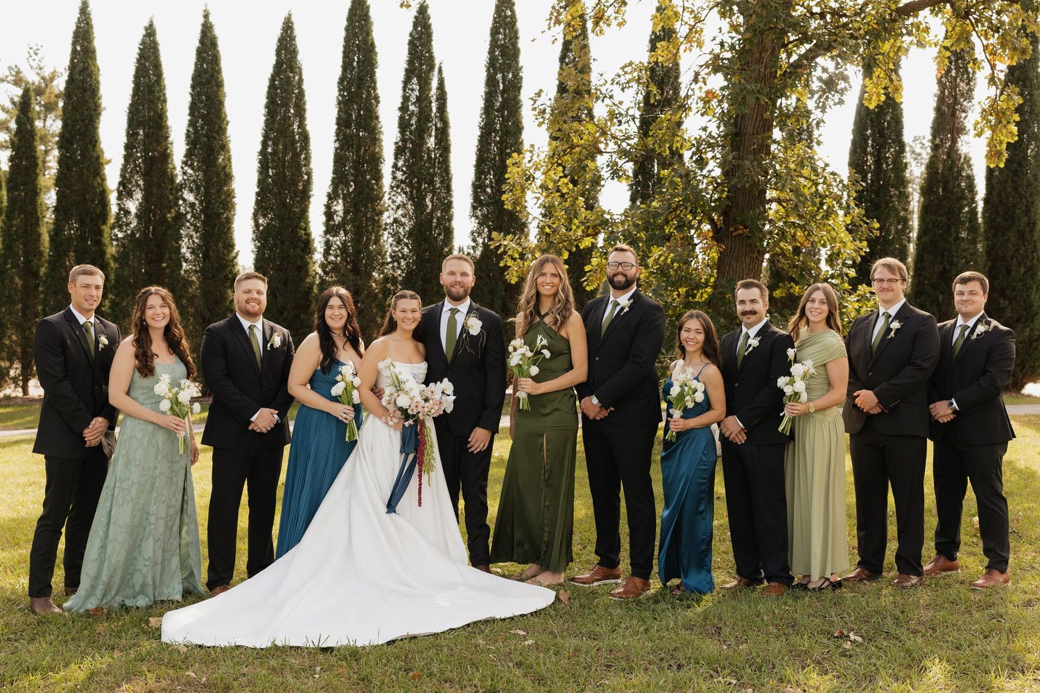 Bride in white gown and groom pose with bridesmaids in green and teal dresses and groomsmen in dark suits among tall cypress trees.