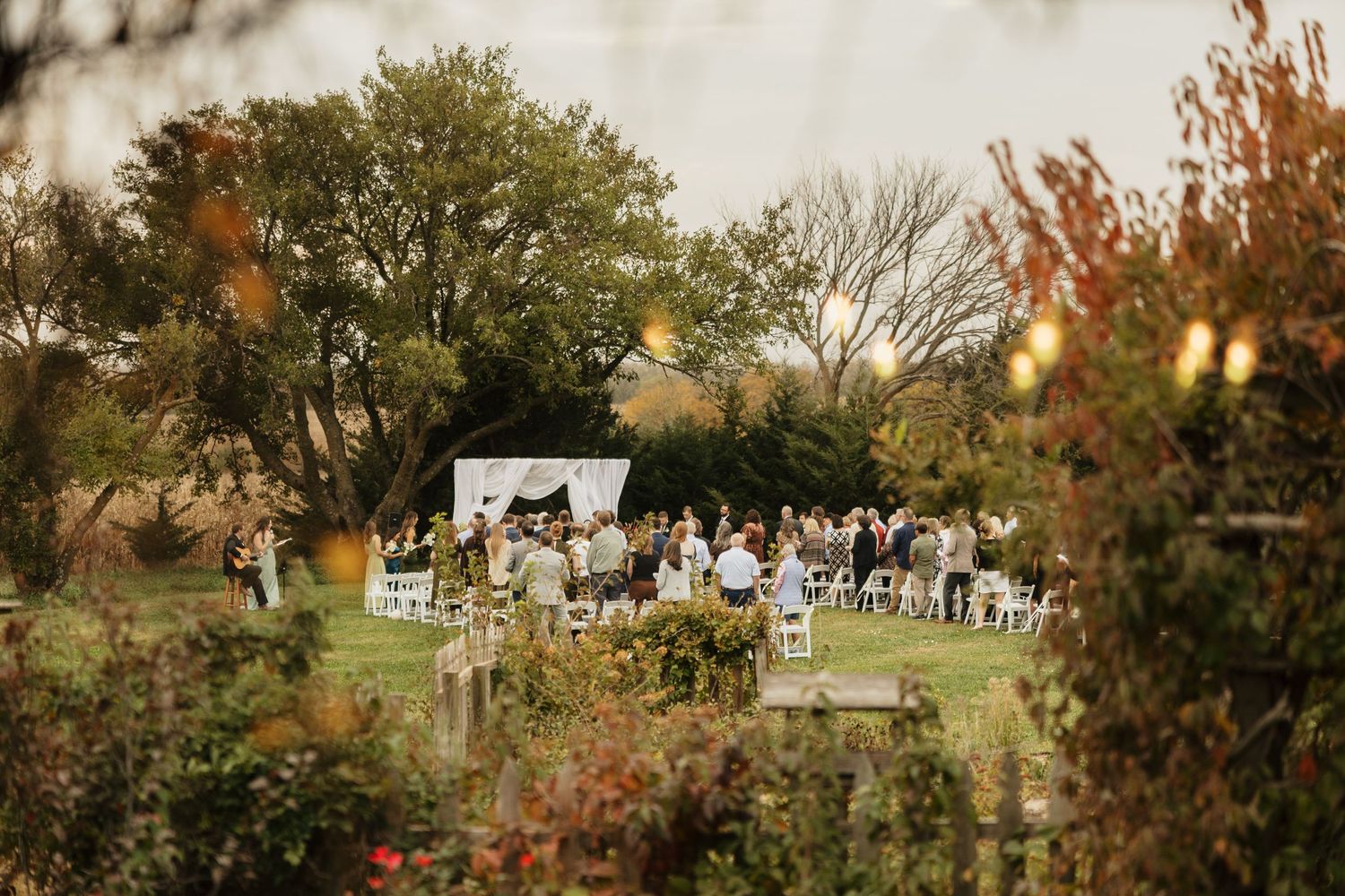 Wedding guests seated on white chairs gather beneath a white canopy on a grassy lawn, framed by autumn foliage in warm evening light.