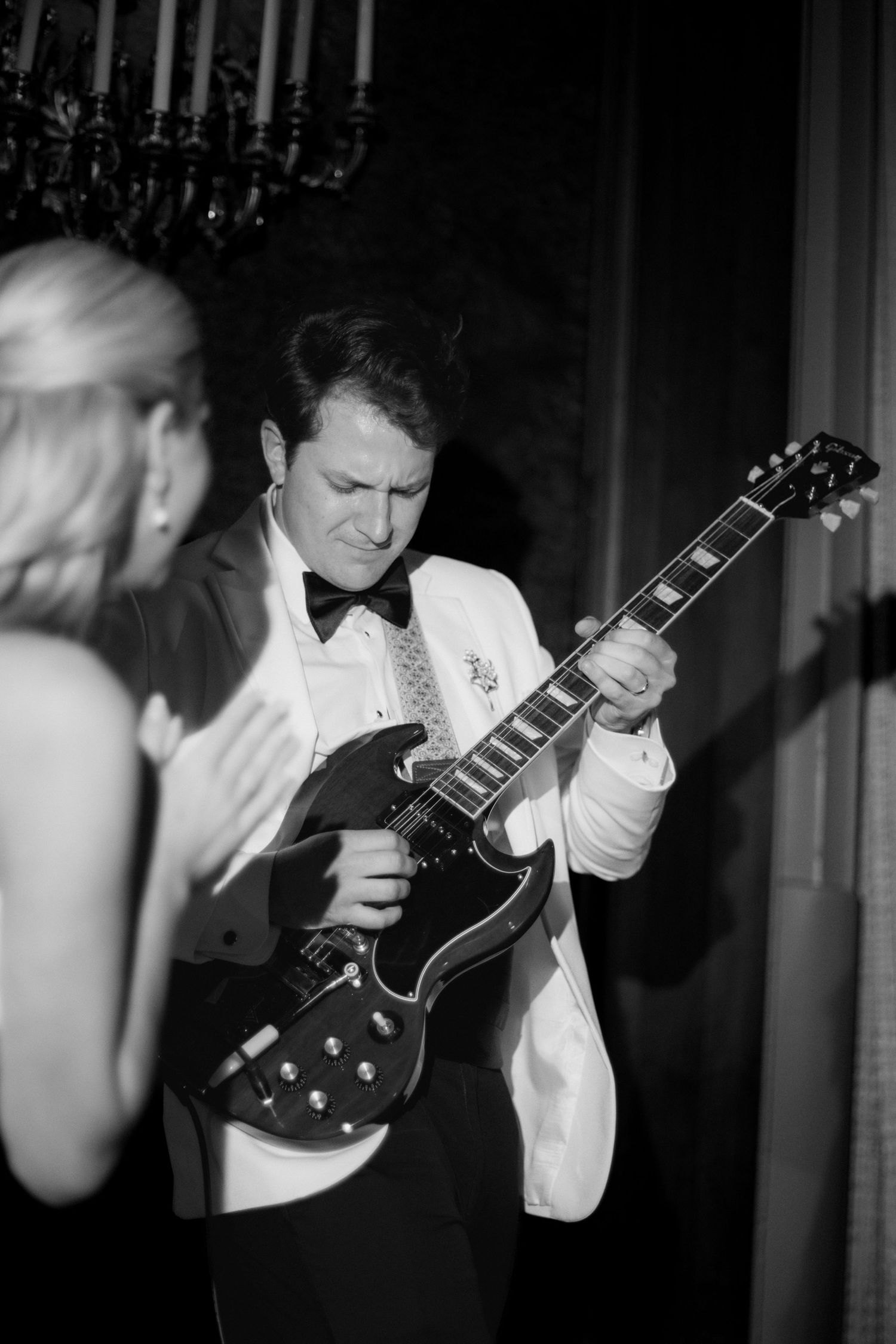 Man in a white dinner jacket and bow tie plays an electric guitar at a formal event while a woman in the foreground applauds.