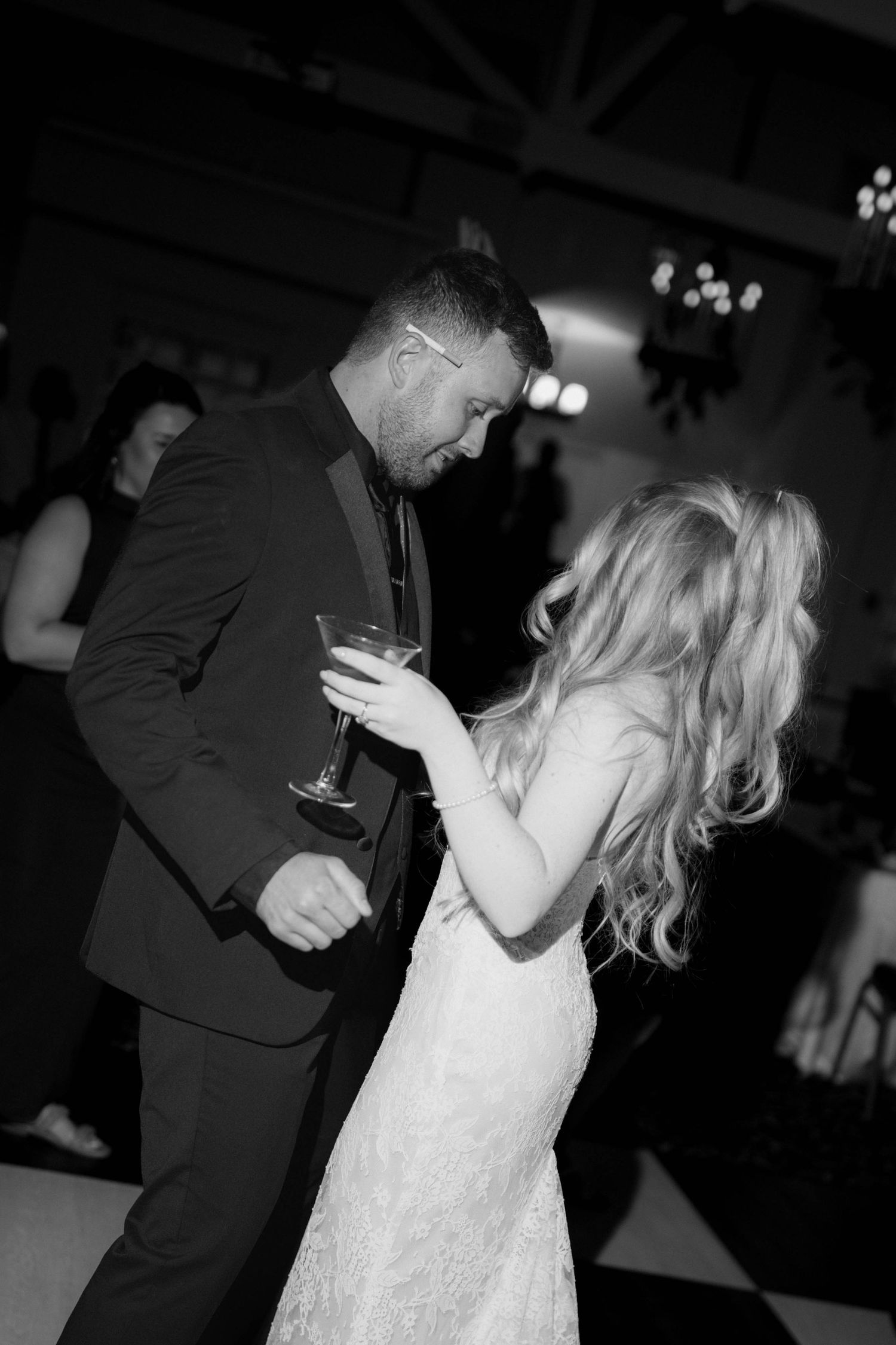 Black and white photo of a groom in a dark suit dancing with a bride in a lace gown, holding a martini glass at a wedding reception.