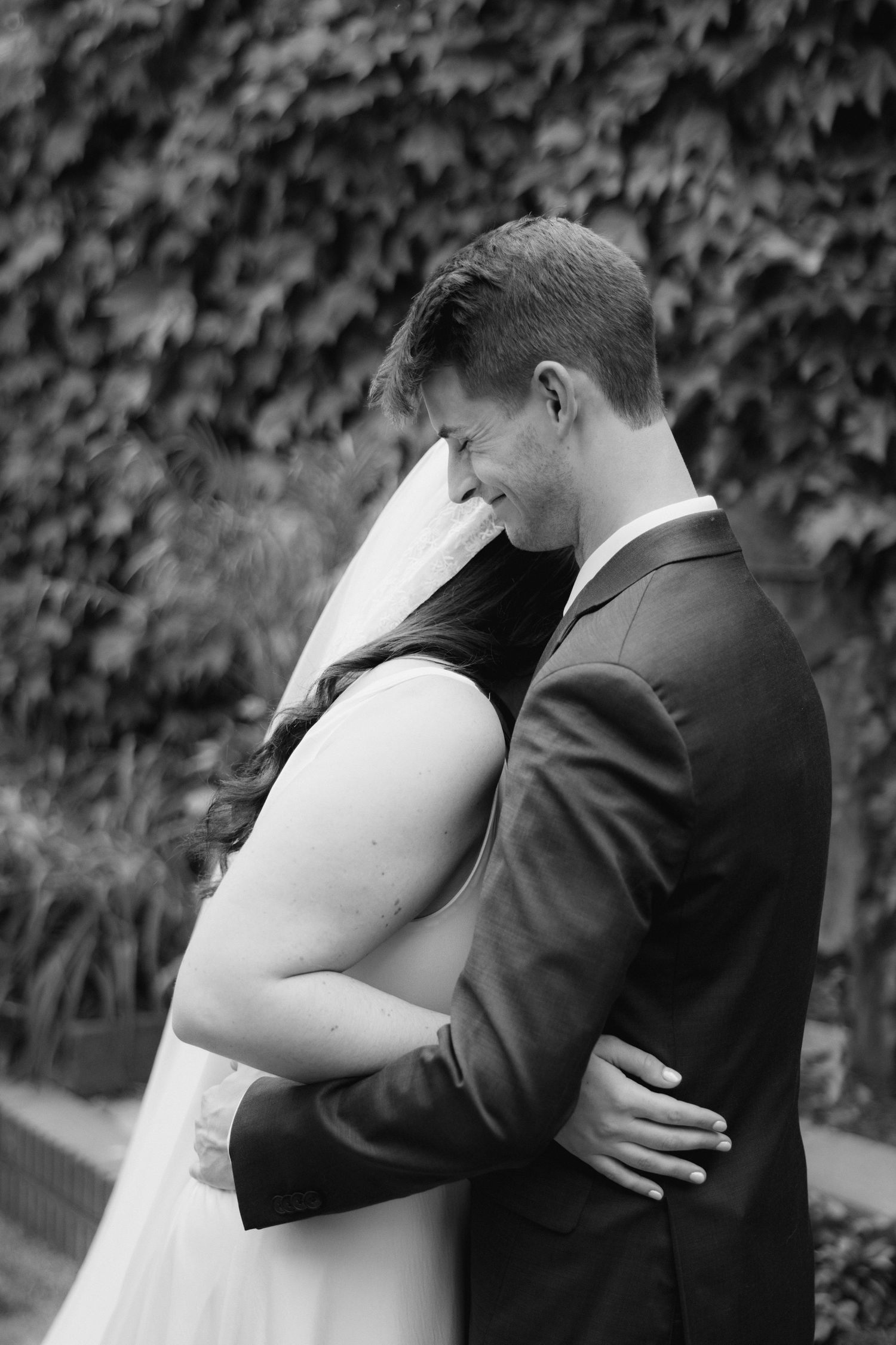 Black and white photo of a smiling groom in a suit embracing his bride in a wedding dress outdoors.