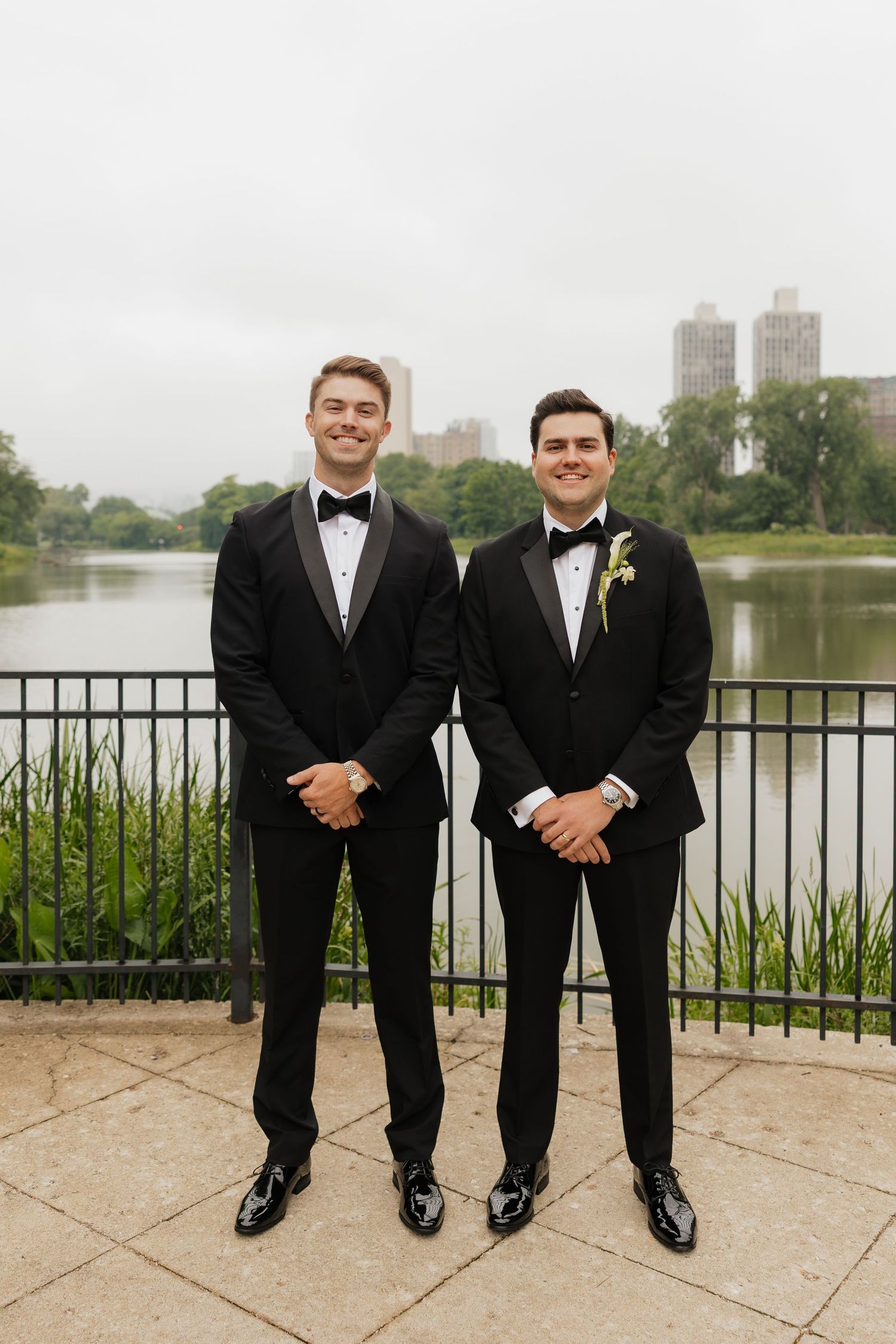 Two grooms in black tuxedos smile outdoors near a lakeside railing with city skyline and greenery behind them.
