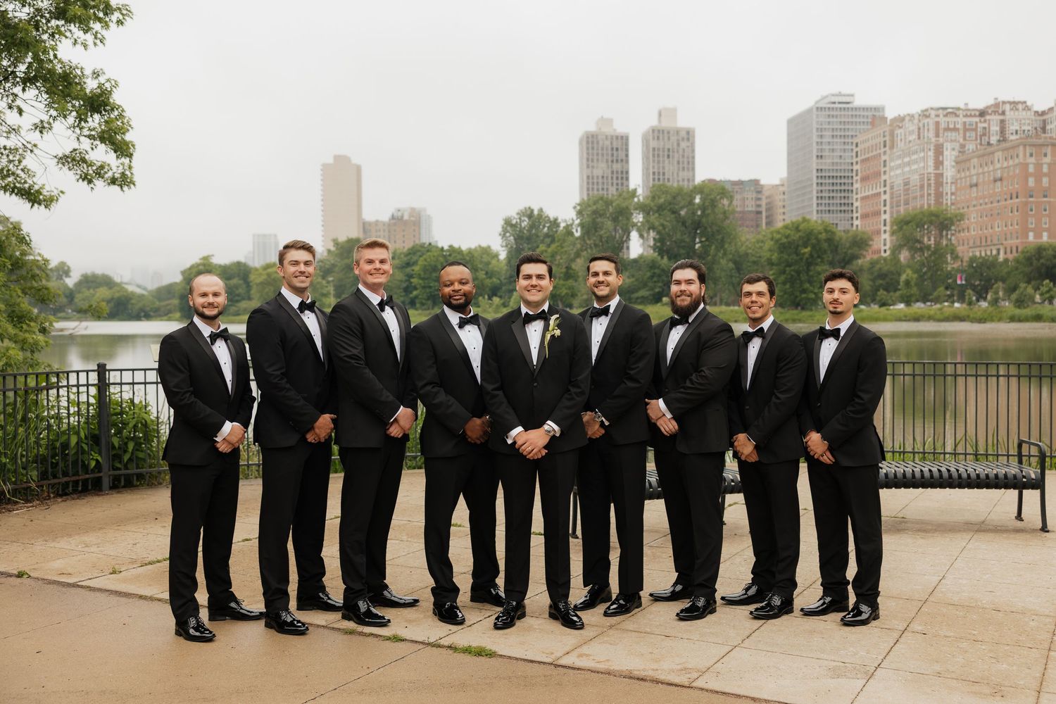 Groom and groomsmen in black tuxedos posing outdoors with Chicago skyline and lake in the background.