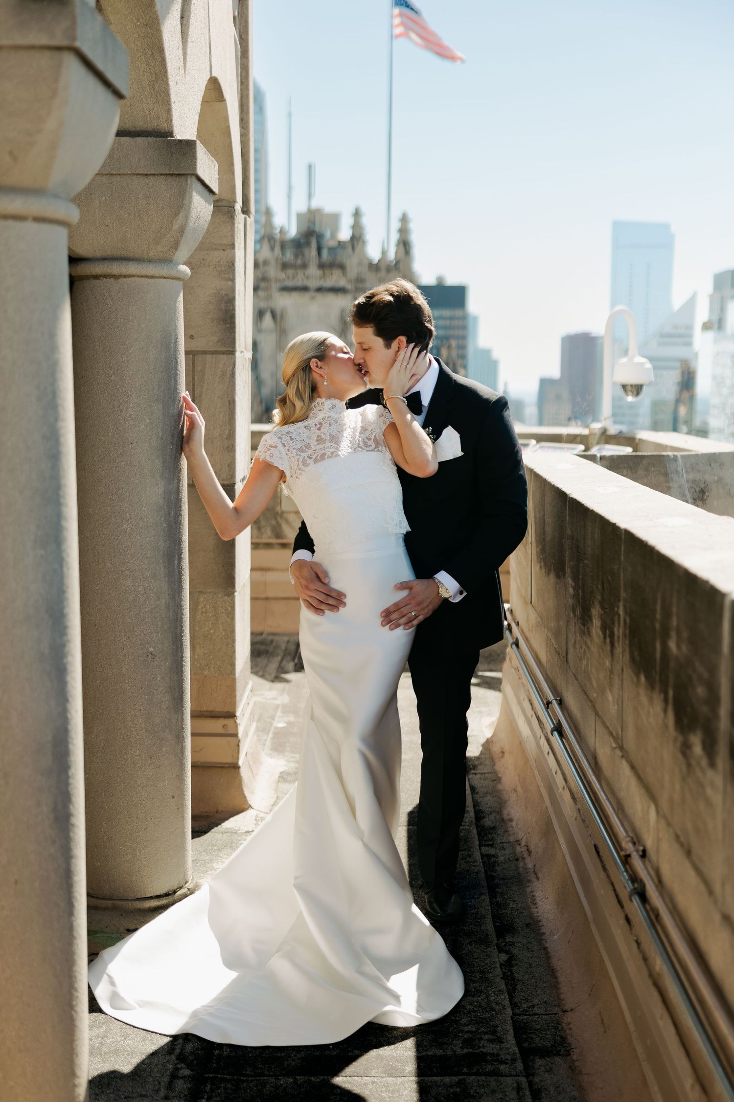 Bride and groom share a romantic kiss on a rooftop terrace with a city skyline backdrop on their wedding day.