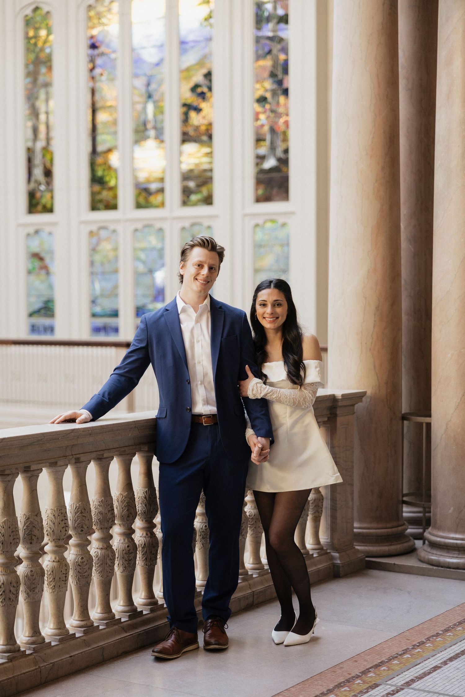A couple poses against classical columns with stained glass windows in the background.