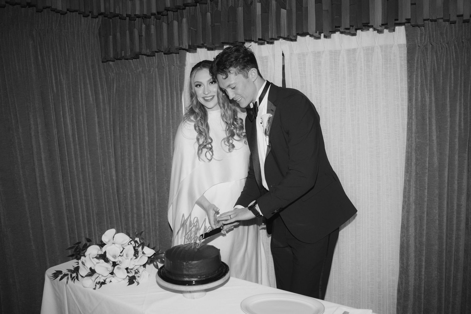 A couple in formal wedding attire cutting their wedding cake together in a black and white photograph.