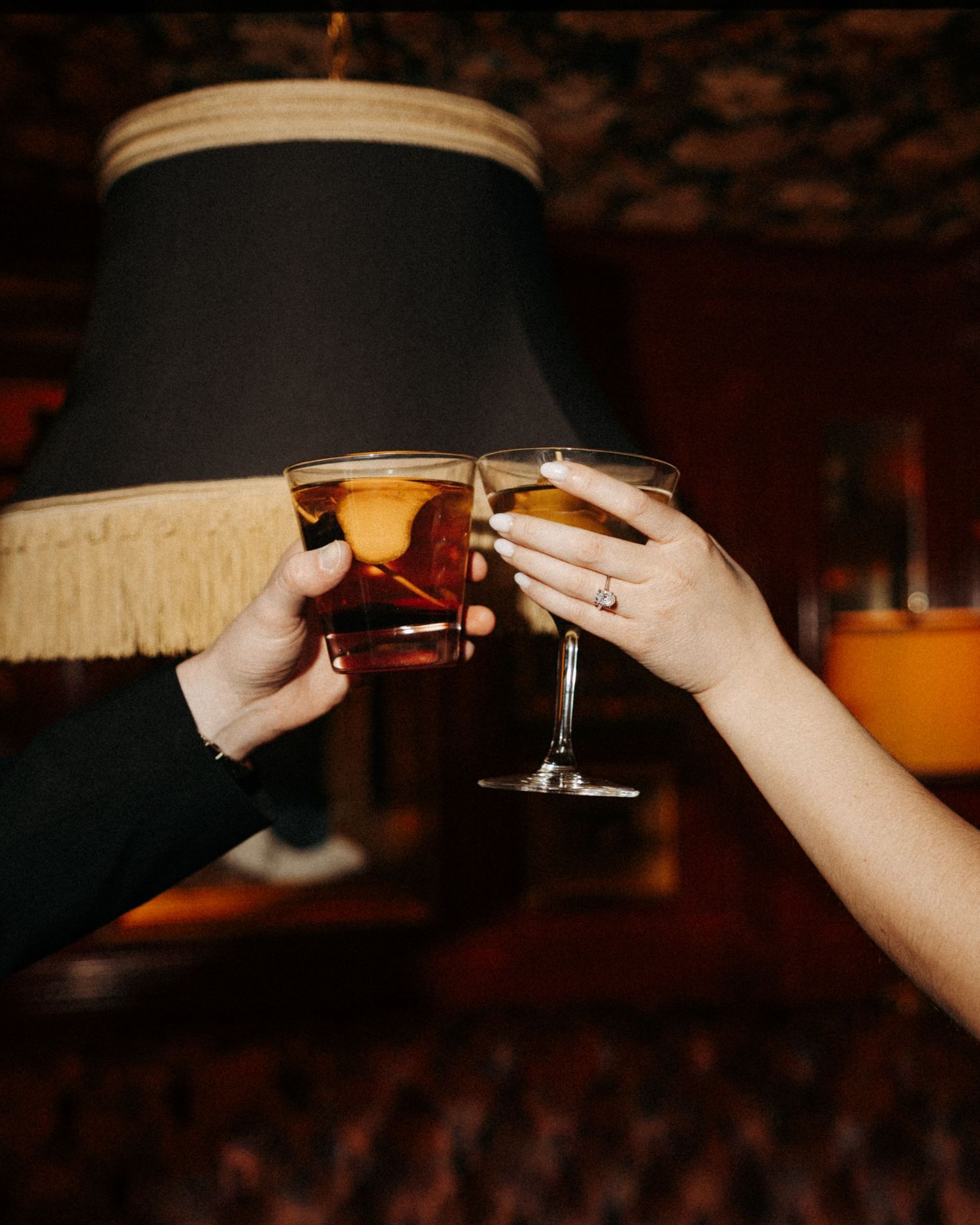 Two hands toasting with cocktail glasses in a dimly lit bar setting with a vintage lamp in the background.