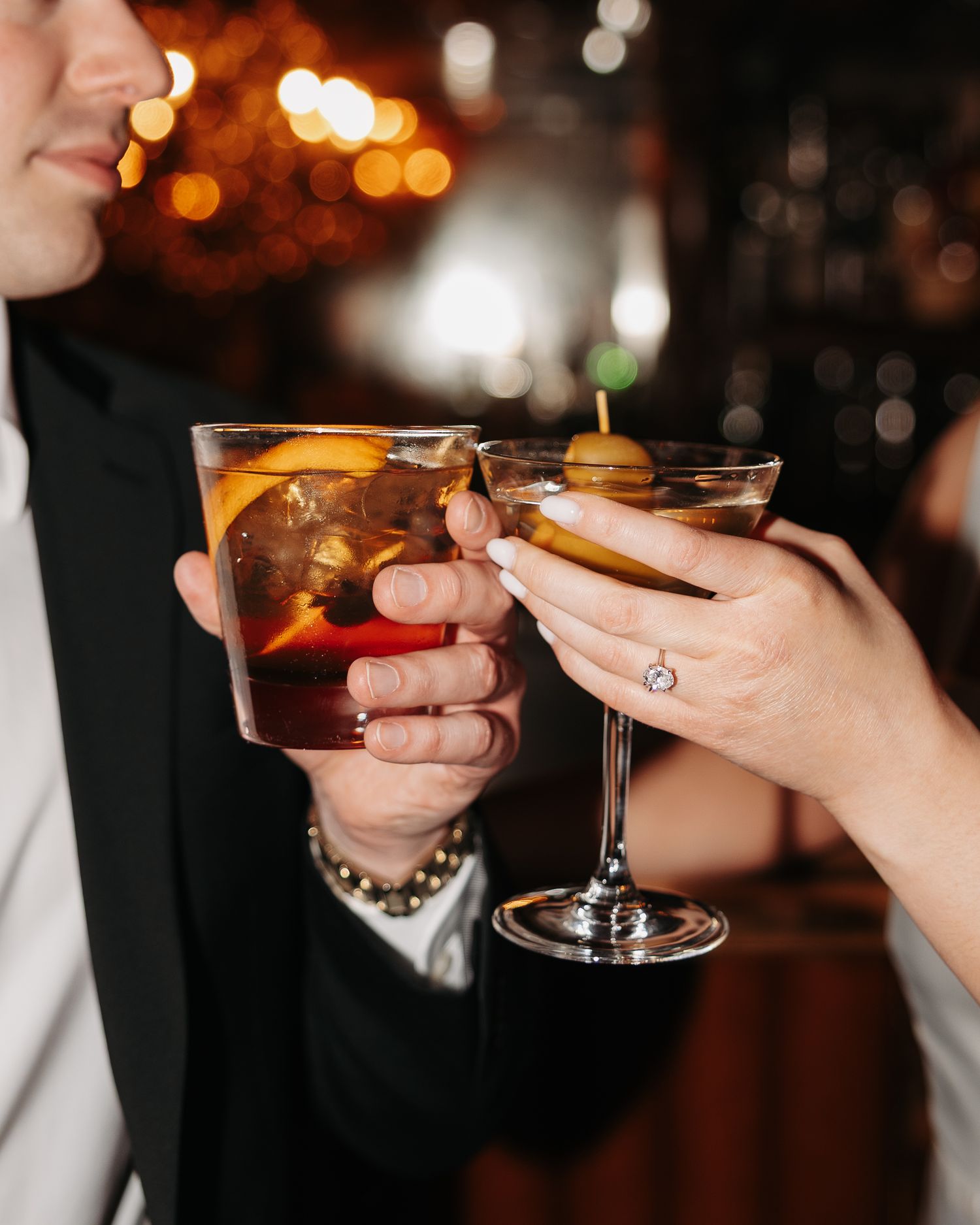 Multiple hands holding cocktail glasses in a toast against a warm, blurred background with bokeh lights.
