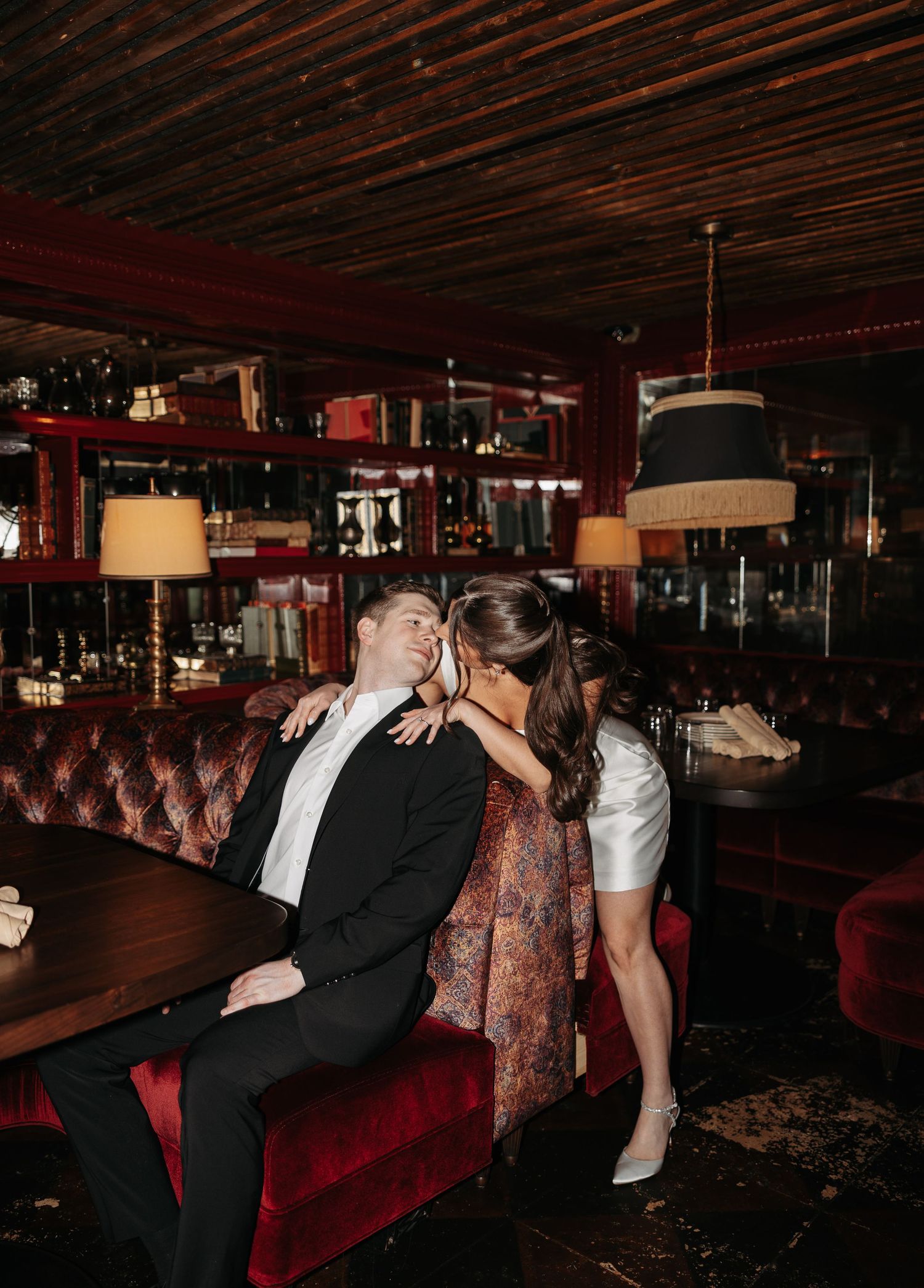A romantic moment captured at a dimly lit vintage bar with red velvet seating and warm lamp lighting.