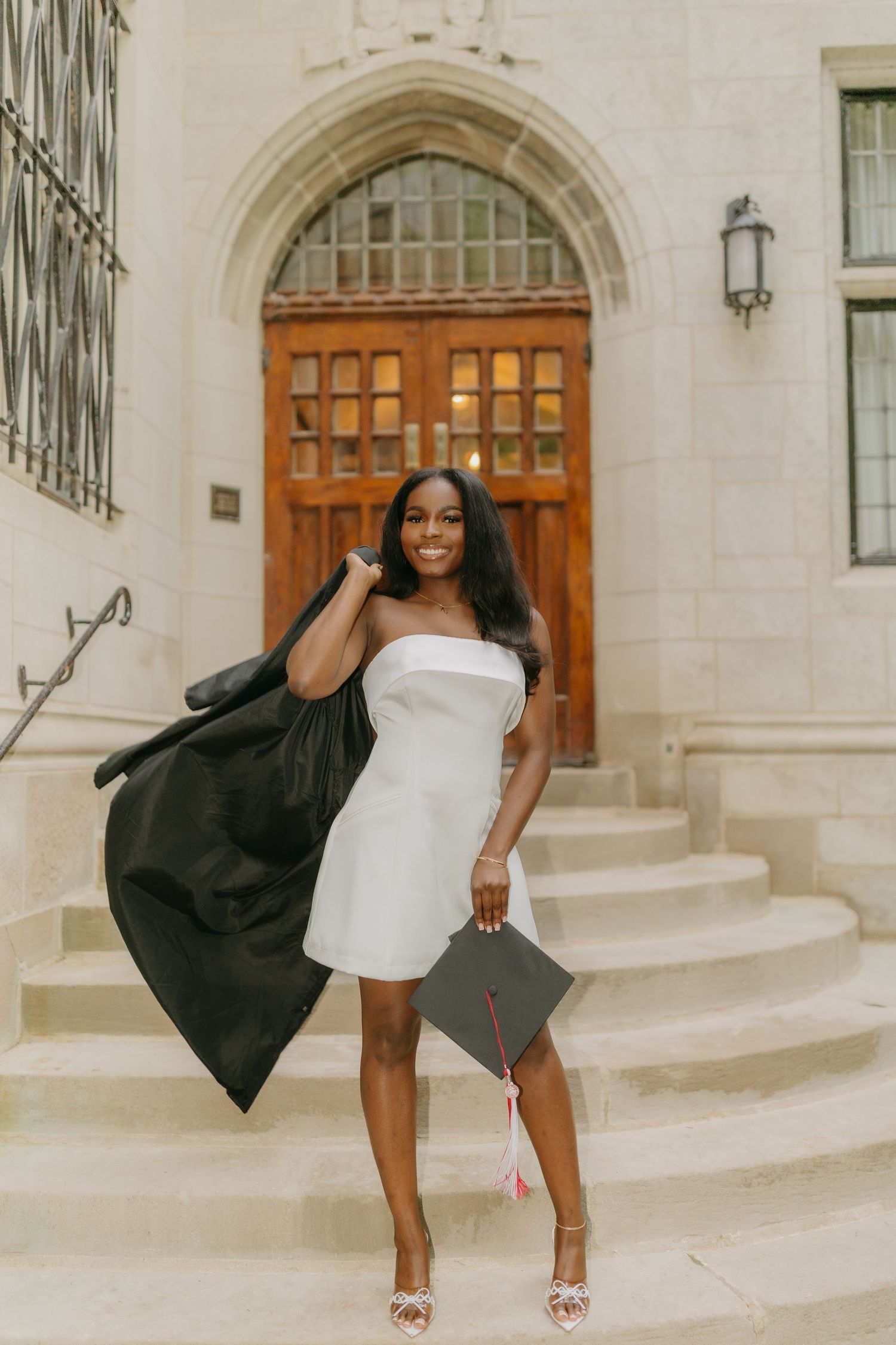A person in a white dress and black jacket poses playfully on stone steps in front of elegant wooden doors.