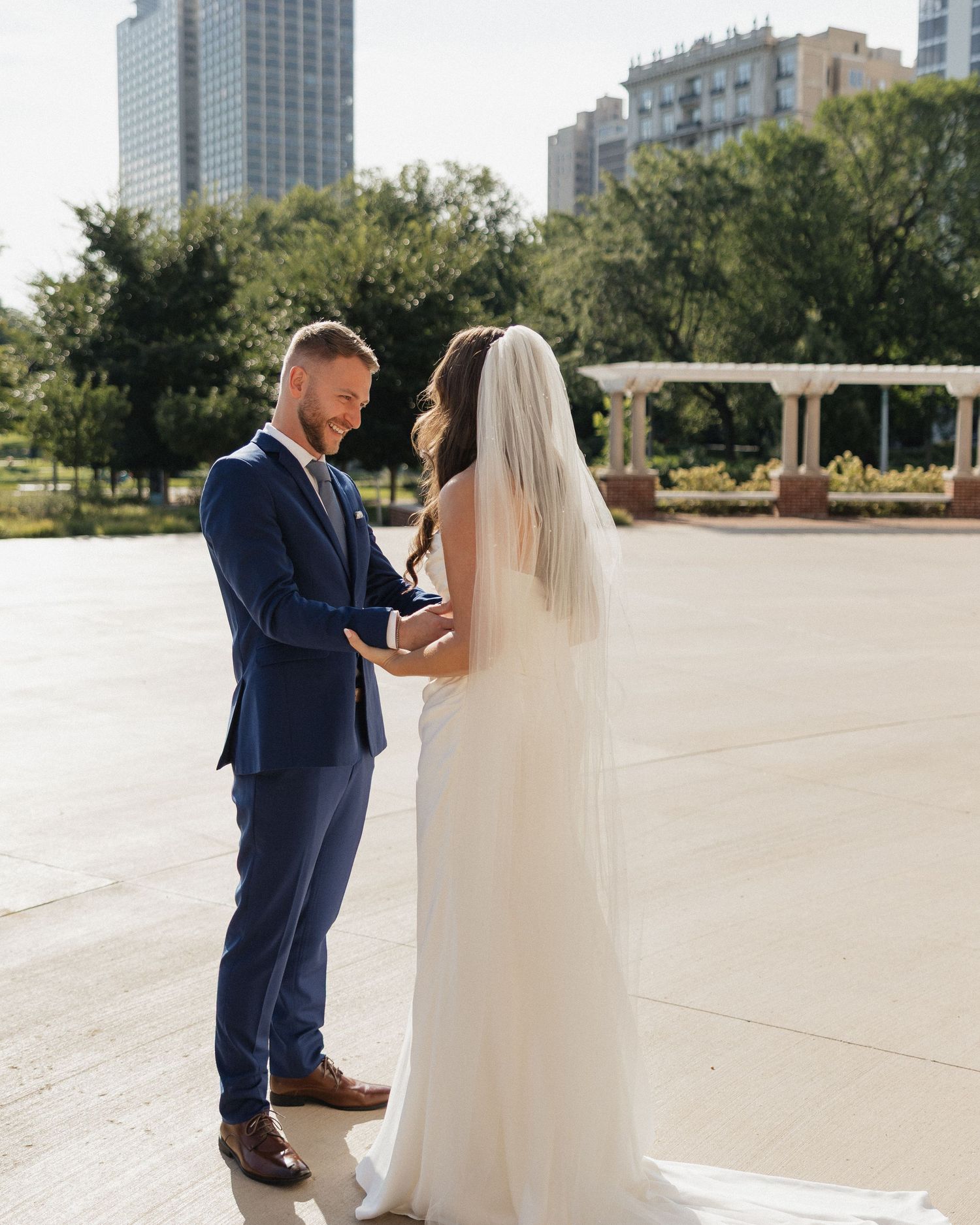 A couple in wedding attire share a romantic moment in an outdoor urban setting with modern buildings in the background.