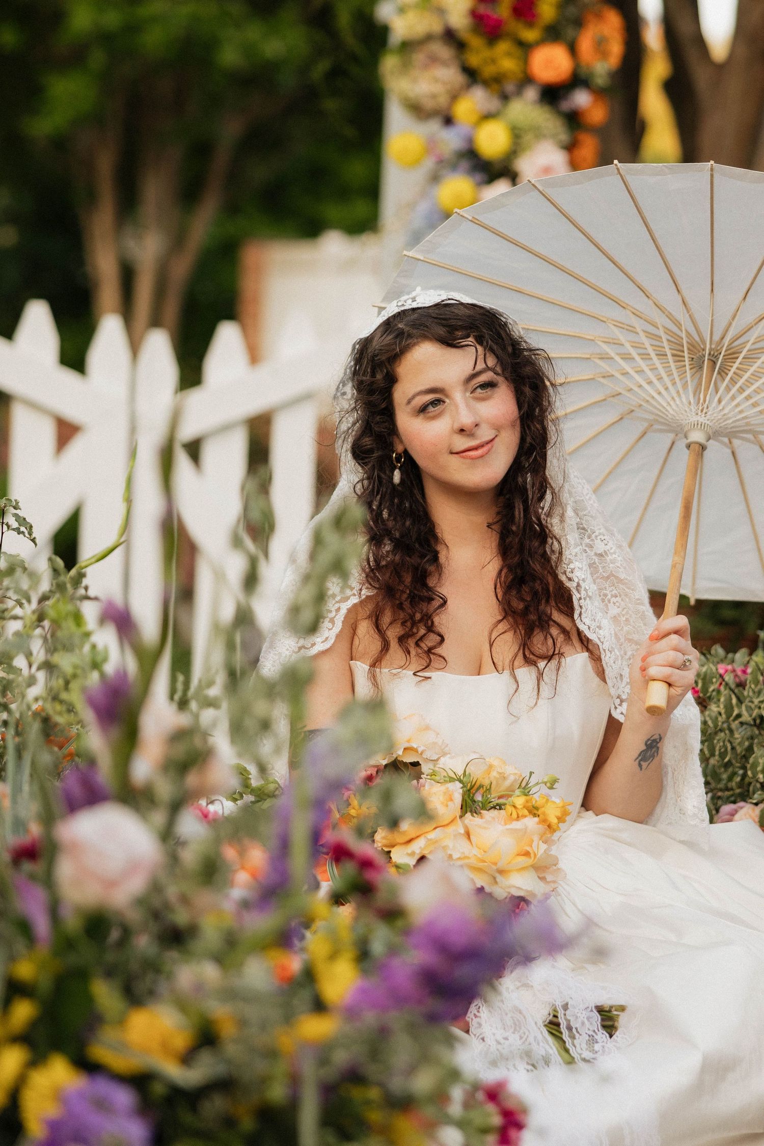 A bride in a white gown holds a parasol and floral bouquet, seated among colorful flowers outdoors.