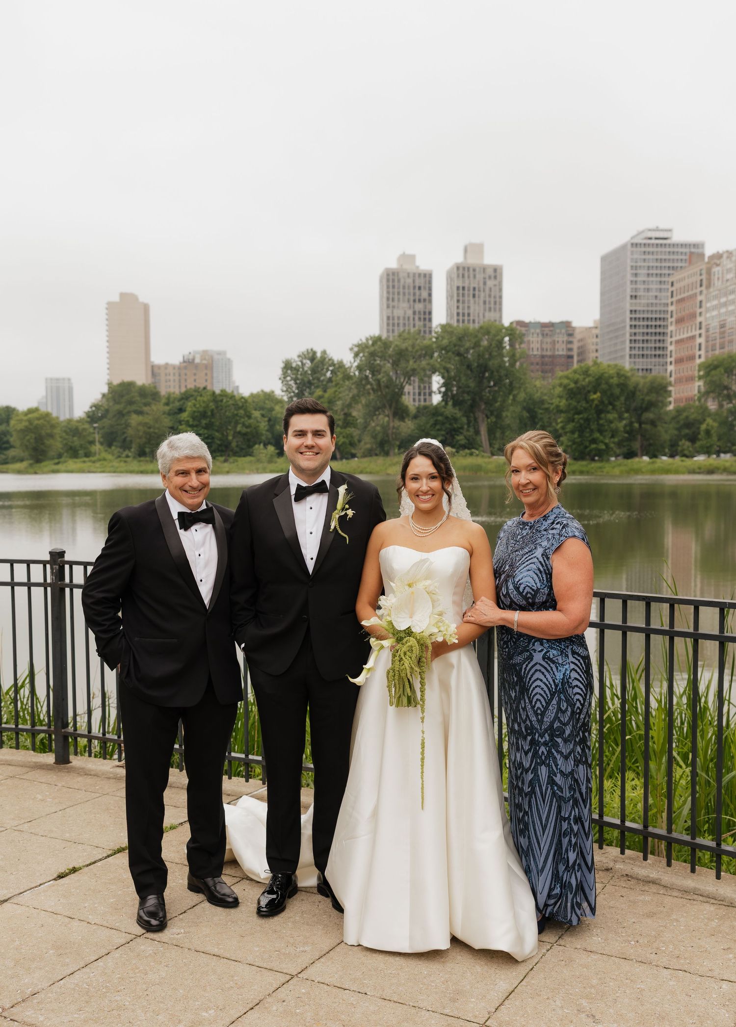 Wedding party of four posing by a lake with city skyline backdrop, bride in white gown holding bouquet.