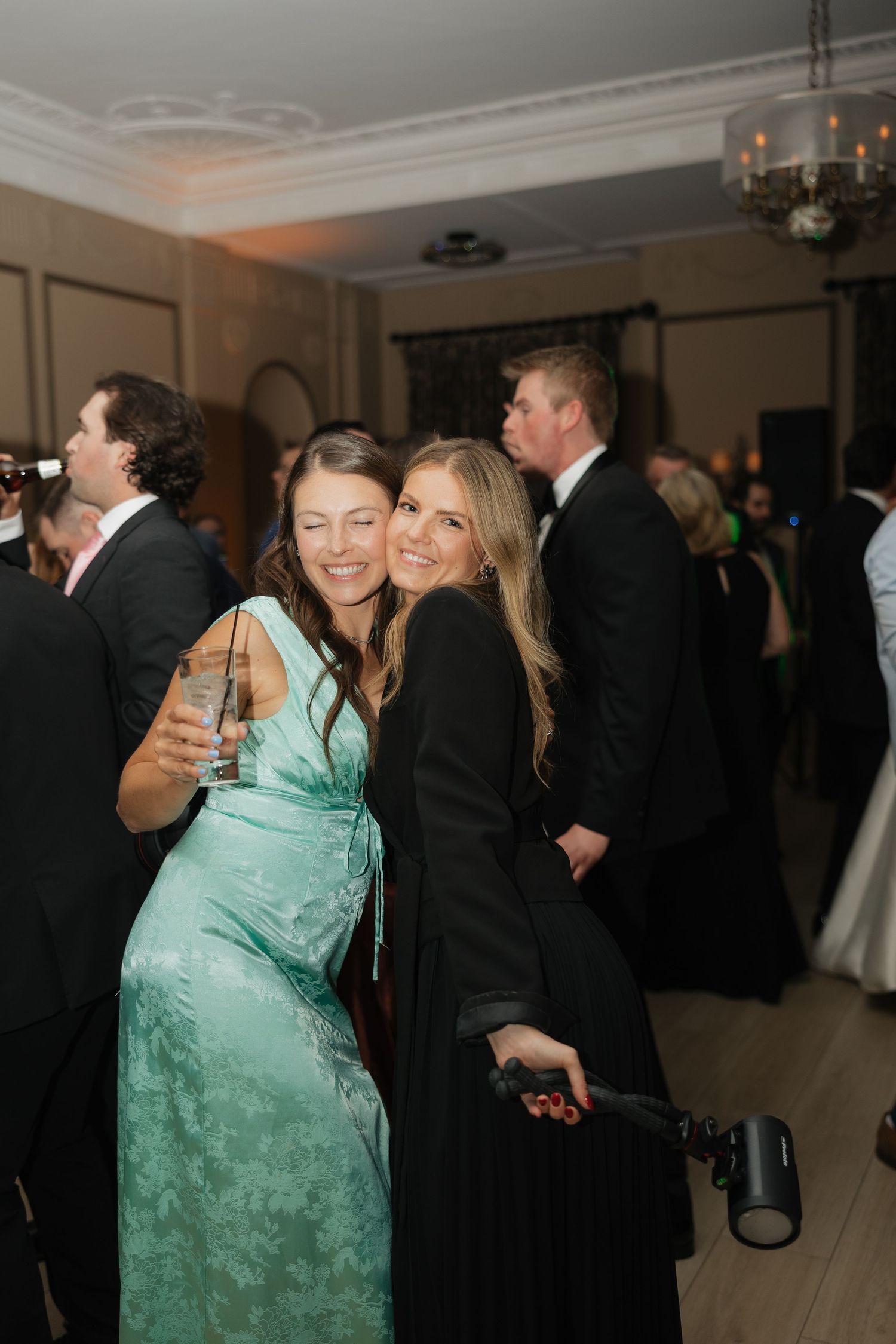 Two women in elegant gowns smiling at a formal event, one in mint green dress holding champagne glass.