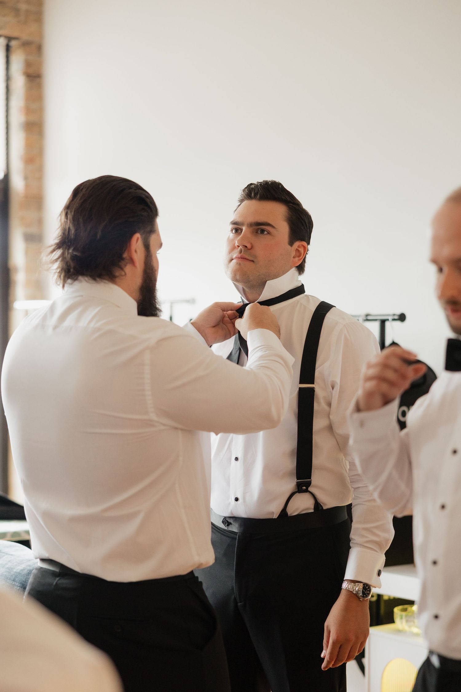 Groomsmen helping groom adjust black bow tie and suspenders while getting ready for wedding ceremony.