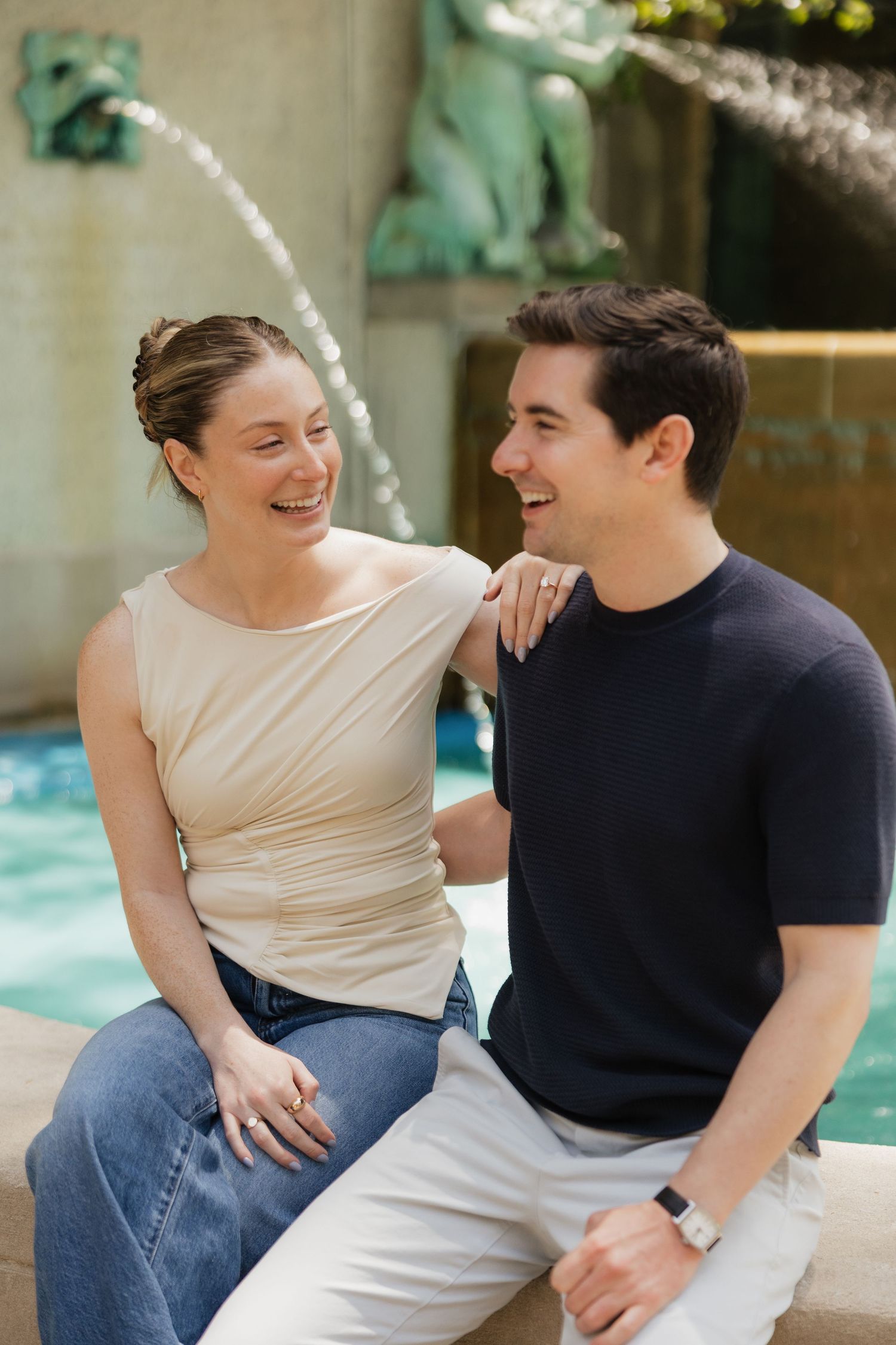 A smiling couple sits together by a decorative fountain, laughing and enjoying each other's company outdoors.