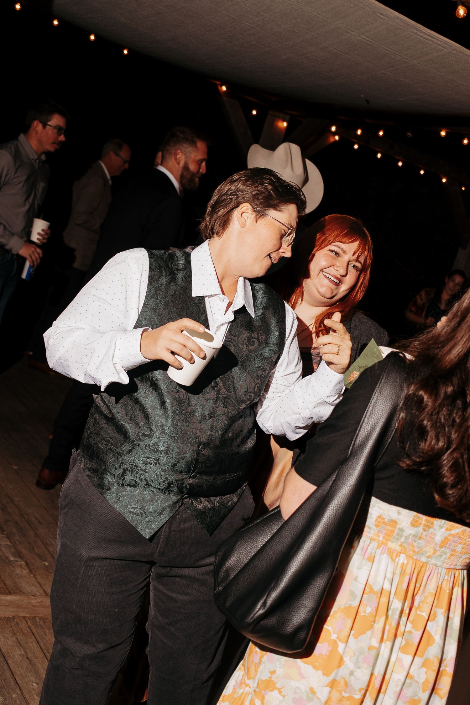 Guests dance and laugh at an evening outdoor party under string lights, one in a green vest holding a cup, another pointing at the camera.