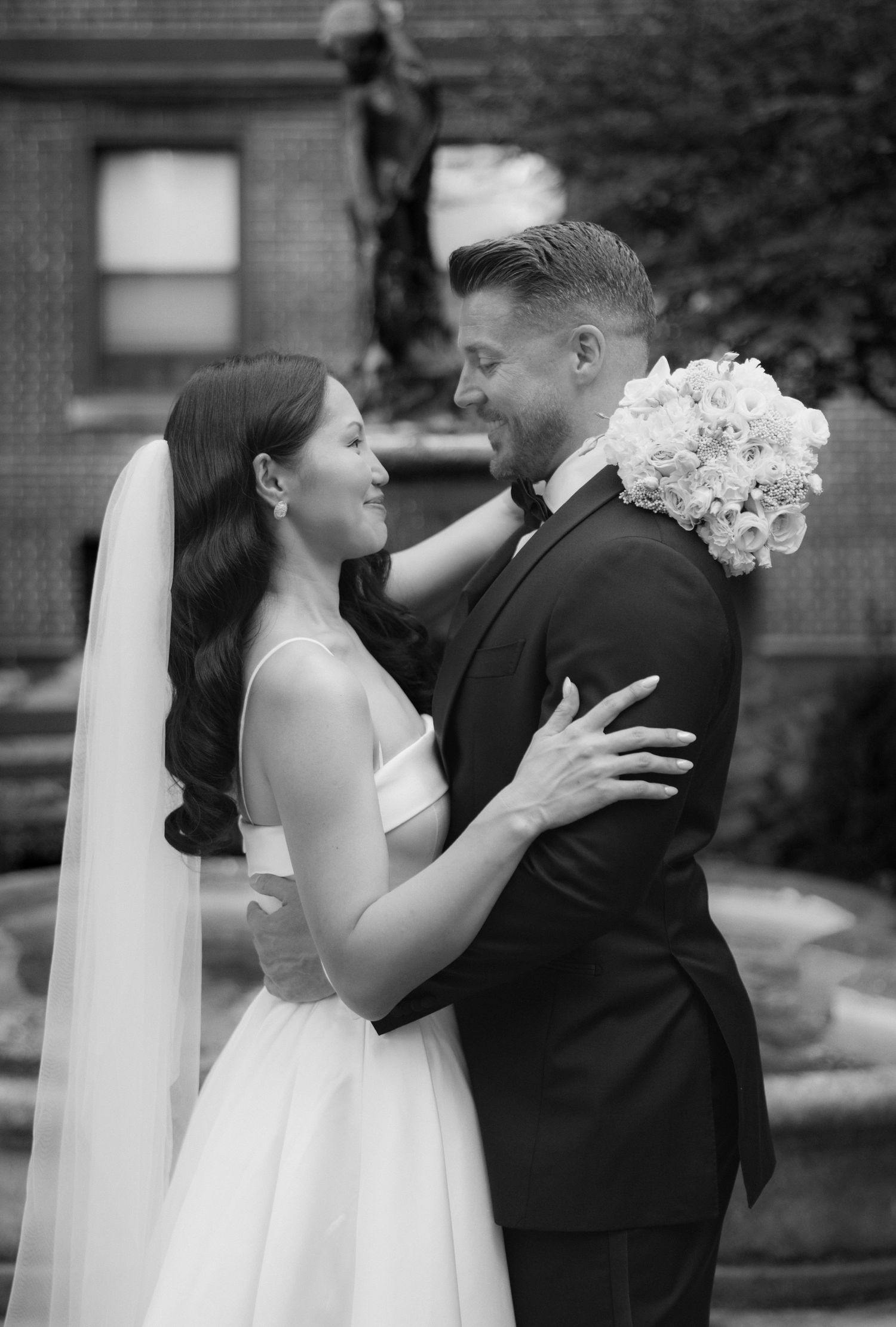 Black and white photo of a bride in a satin gown and veil smiling at her groom in a tuxedo holding a white floral bouquet near a fountain.