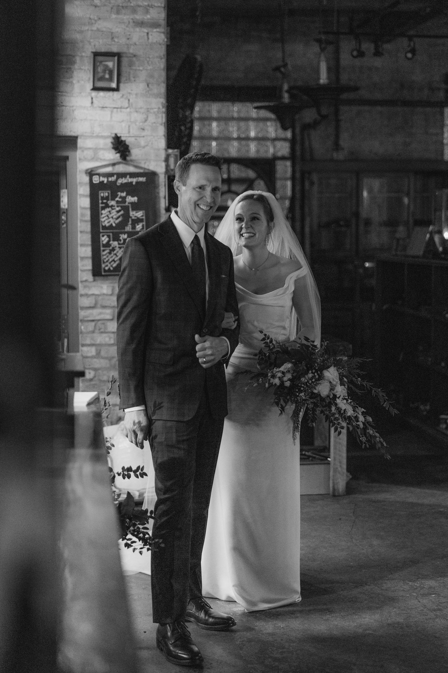 A couple in wedding attire shares an intimate moment in a rustic brick-walled venue with soft lighting.