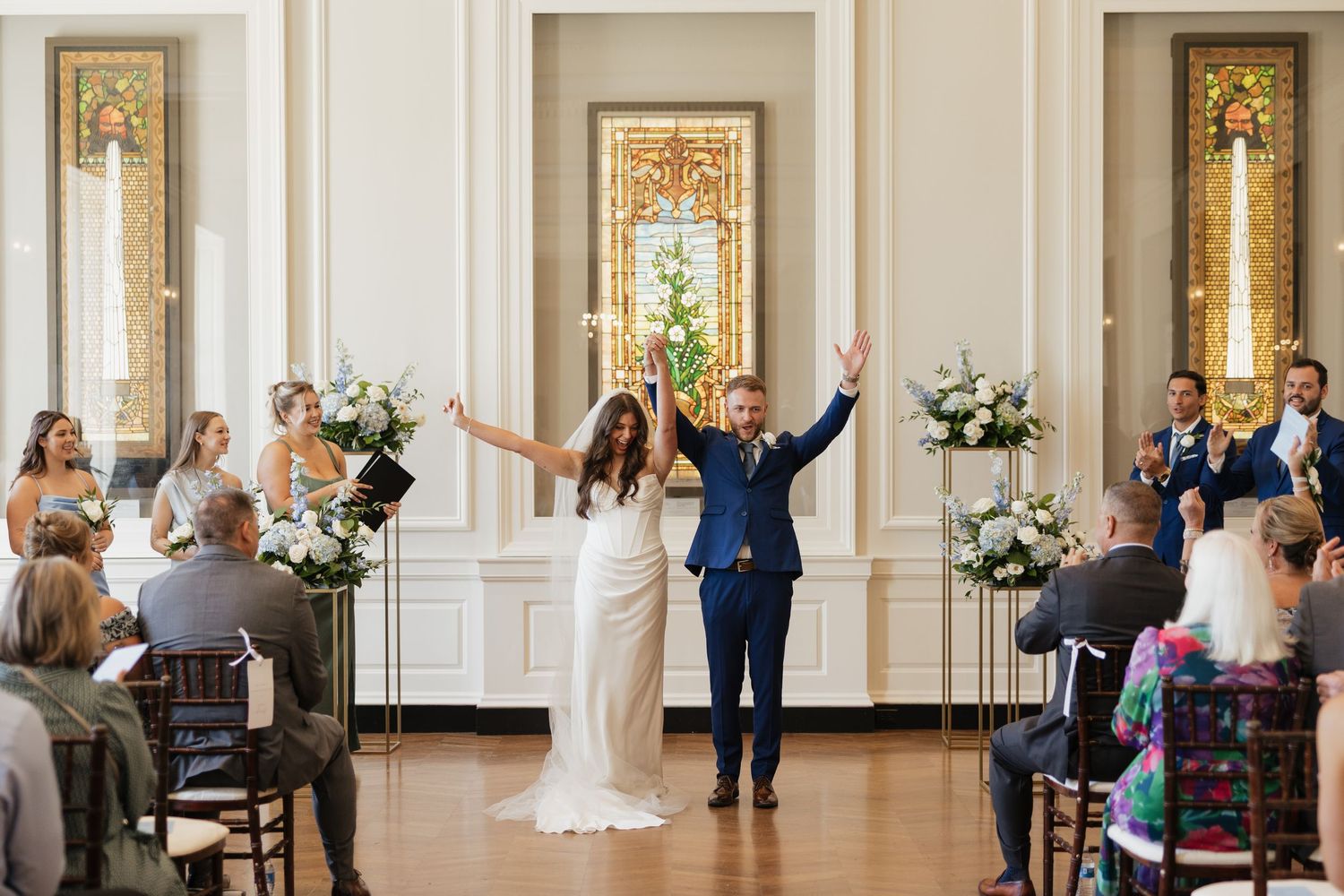 Elegant wedding party dances and celebrates in a grand white ballroom with ornate gold details and floral arrangements.