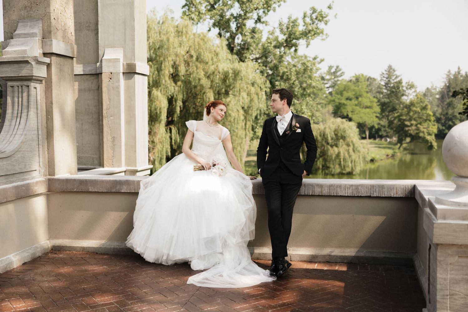 Bride in white ballgown and groom in black tuxedo pose on stone terrace with willow tree and pond backdrop.