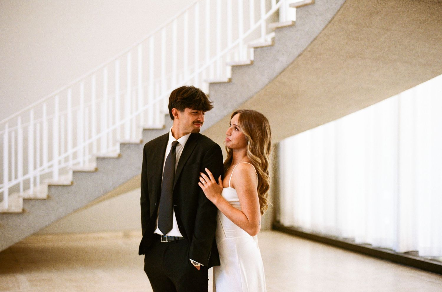 Couple in formal attire sharing a romantic moment near a spiral staircase in an elegant venue.