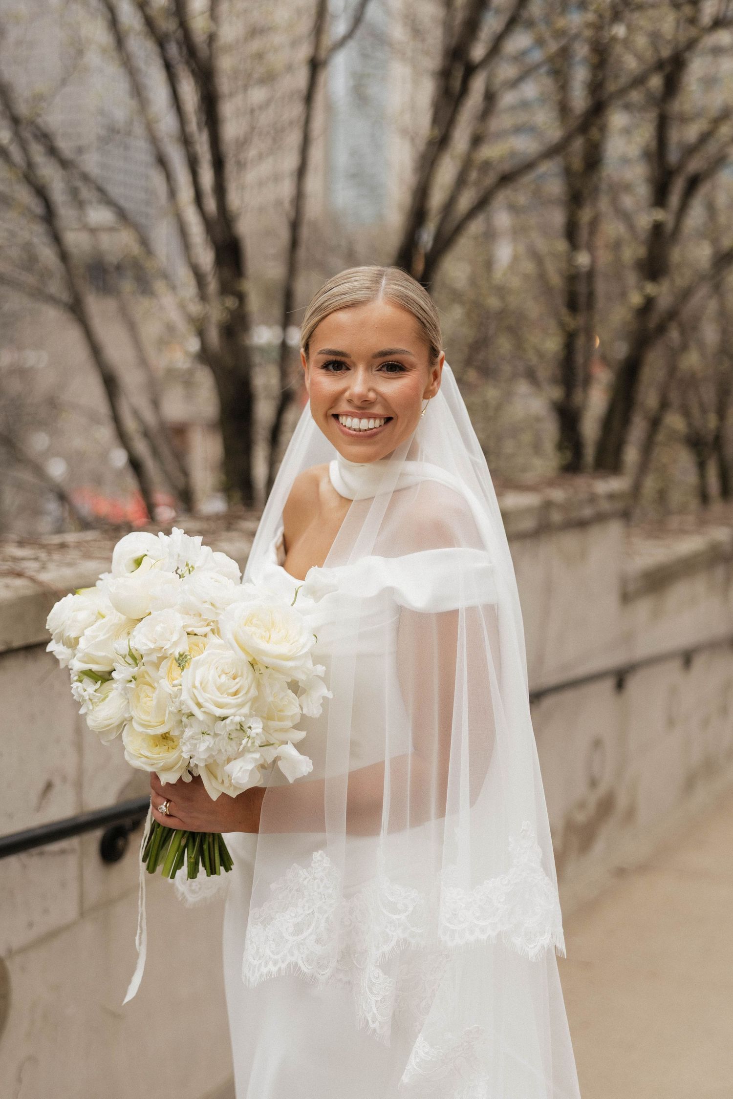 Smiling bride in white lace gown and veil holds white rose bouquet outdoors in urban setting.