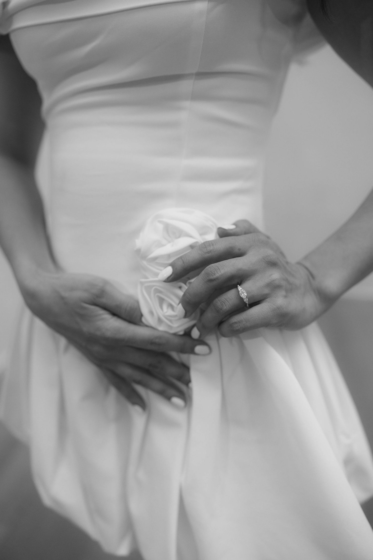 Close-up black and white photo of bride's hands holding white rose detail on wedding dress with ring.