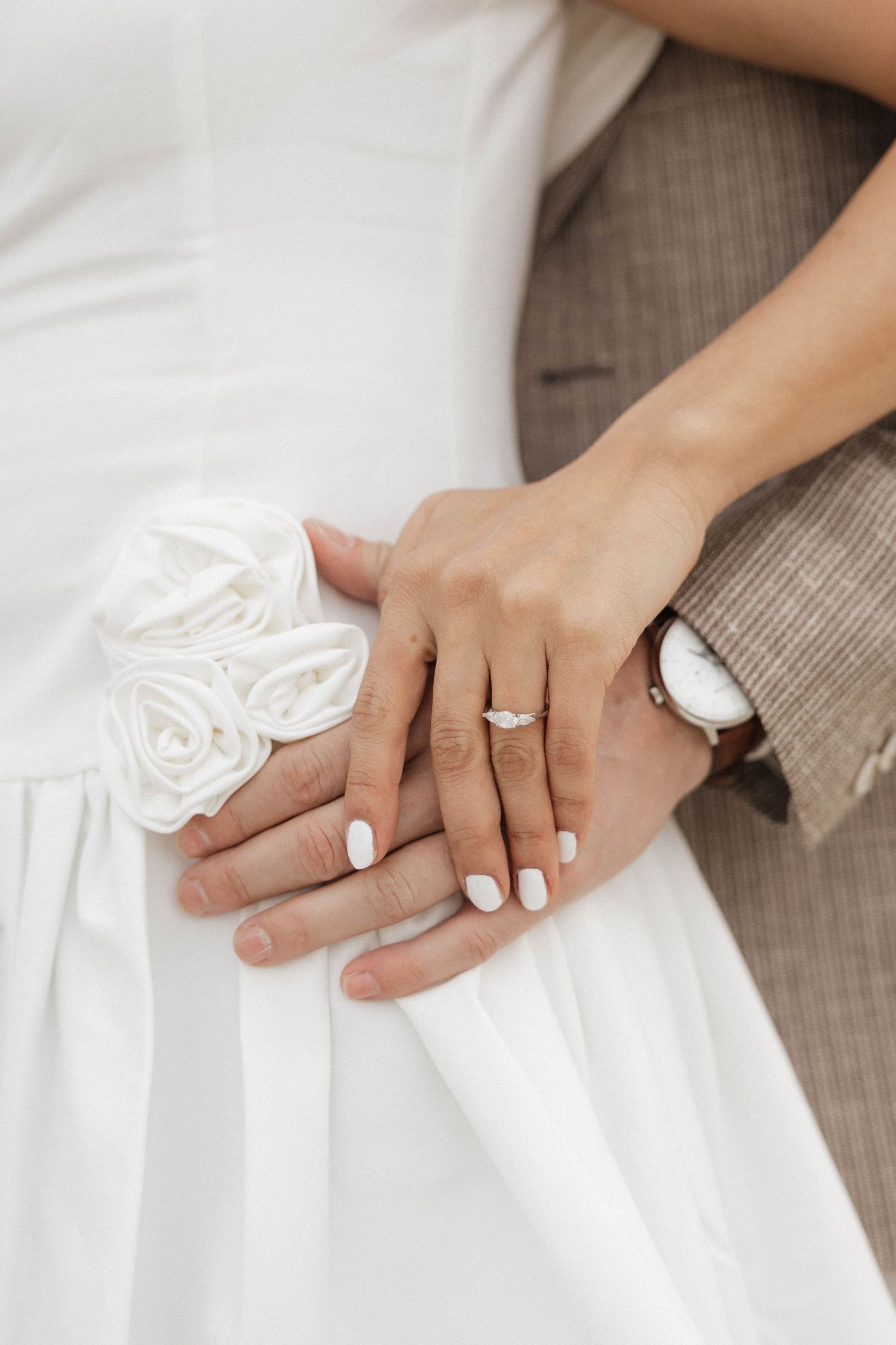 Close-up of bride and groom hands showing engagement ring on white wedding dress with floral detail.